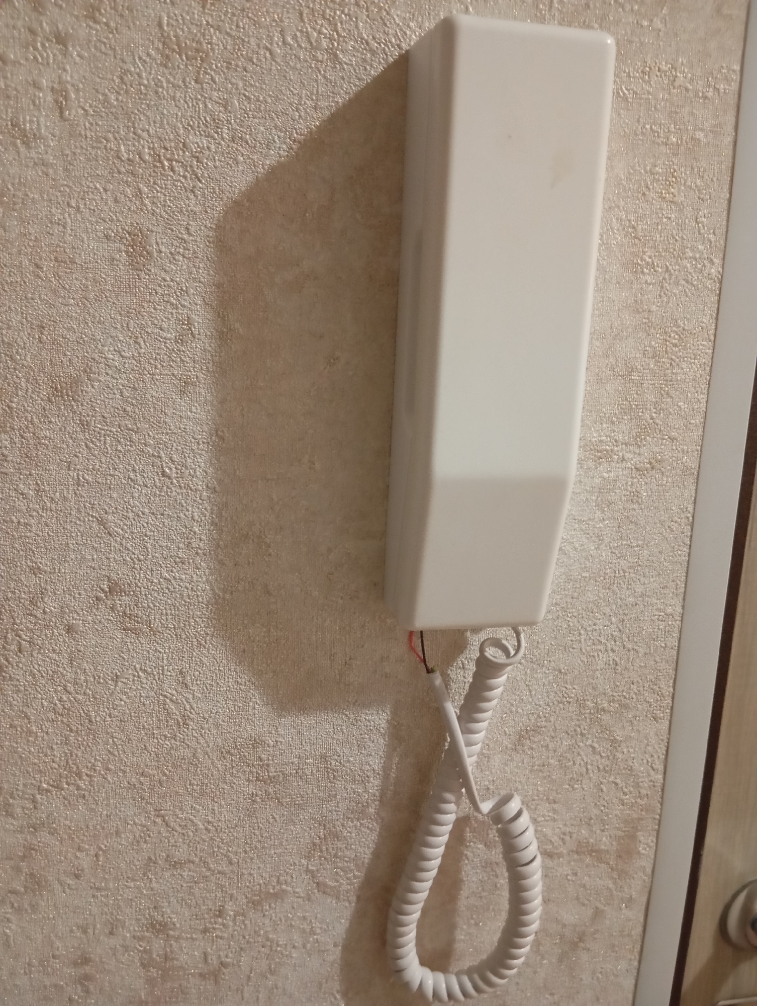 A white intercom unit is mounted on a textured, light-colored wall. The intercom has a coiled white cord hanging from it, which is partially visible. The cord is connected to a handset (not visible in full). The wall has a bumpy, uneven texture, and there is a shadow cast on the wall to the left of the intercom, suggesting a light source from the right. The scene appears to be indoors, possibly in a hallway or near a doorway, as suggested by a vertical white frame on the right side of the image and a hint of a door handle. The lighting is moderate, and the overall image is clear.