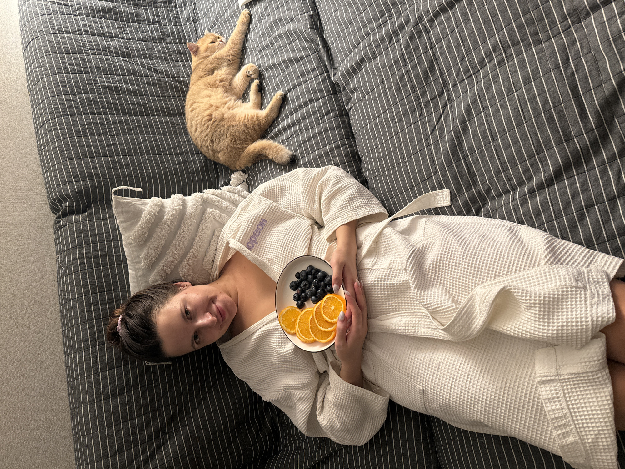 This indoor overhead shot captures a woman and a cat relaxing on a bed, likely in Odintsovo, Russia. The woman is positioned on the lower left side of the frame, lying on her back with her head turned towards the camera, displaying a gentle smile. She wears a white waffle-knit bathrobe, with the text "HOED" visible sideways on its collar. In her hands, she holds a white plate containing dark blueberries and several bright orange slices.
In the upper right section of the bed, a light brown or ginger cat is sprawled on its back or side, with its paws stretched upwards, appearing relaxed.
The bed is covered with dark grey bedding featuring narrow white stripes. A white pillow is visible beneath the woman's head. The scene conveys a sense of calm and domestic comfort. The lighting suggests it is daytime.