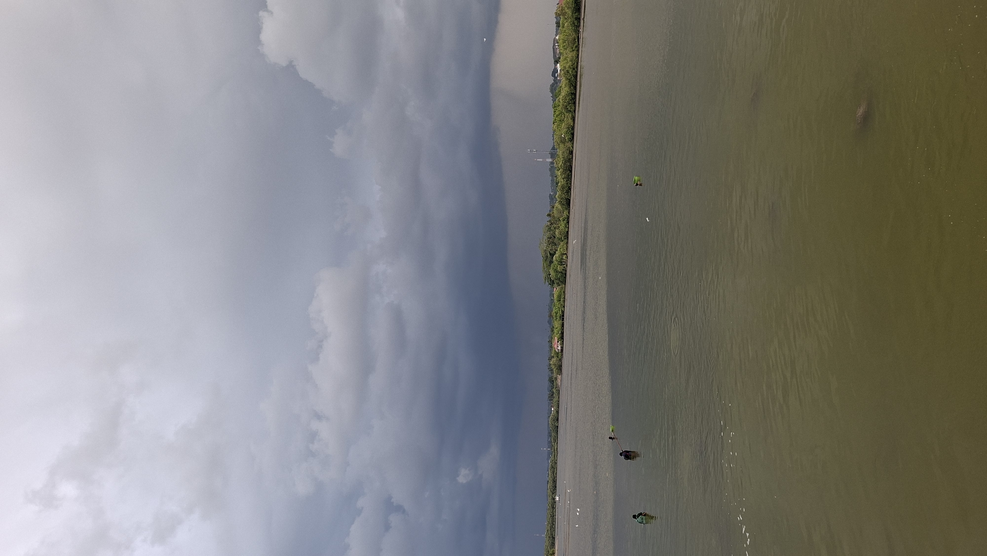 This is a photograph of a body of water, possibly a river or bay, in Lhokseumawe, Indonesia. The sky is overcast with dark, ominous storm clouds, suggesting impending rain. The water is a murky green-brown color, and appears calm with gentle ripples. In the middle distance, along the shoreline, is a dense line of green trees and vegetation. Scattered amongst the trees are some buildings and what appear to be telecommunication towers.
In the water, there are a few individuals. On the left side of the image, near the shore, two people are wading in the water. One person, wearing a green shirt, is facing towards the right, while another person, wearing darker clothing, is facing away from the camera and appears to be holding a fishing net. Further out in the water, closer to the center, is another individual, also partially submerged. There are also a few white birds on the water's surface in the middle distance, and what appears to be a green bucket floating. The overall impression is of a natural landscape under a dramatic sky, with human activity present in the water.