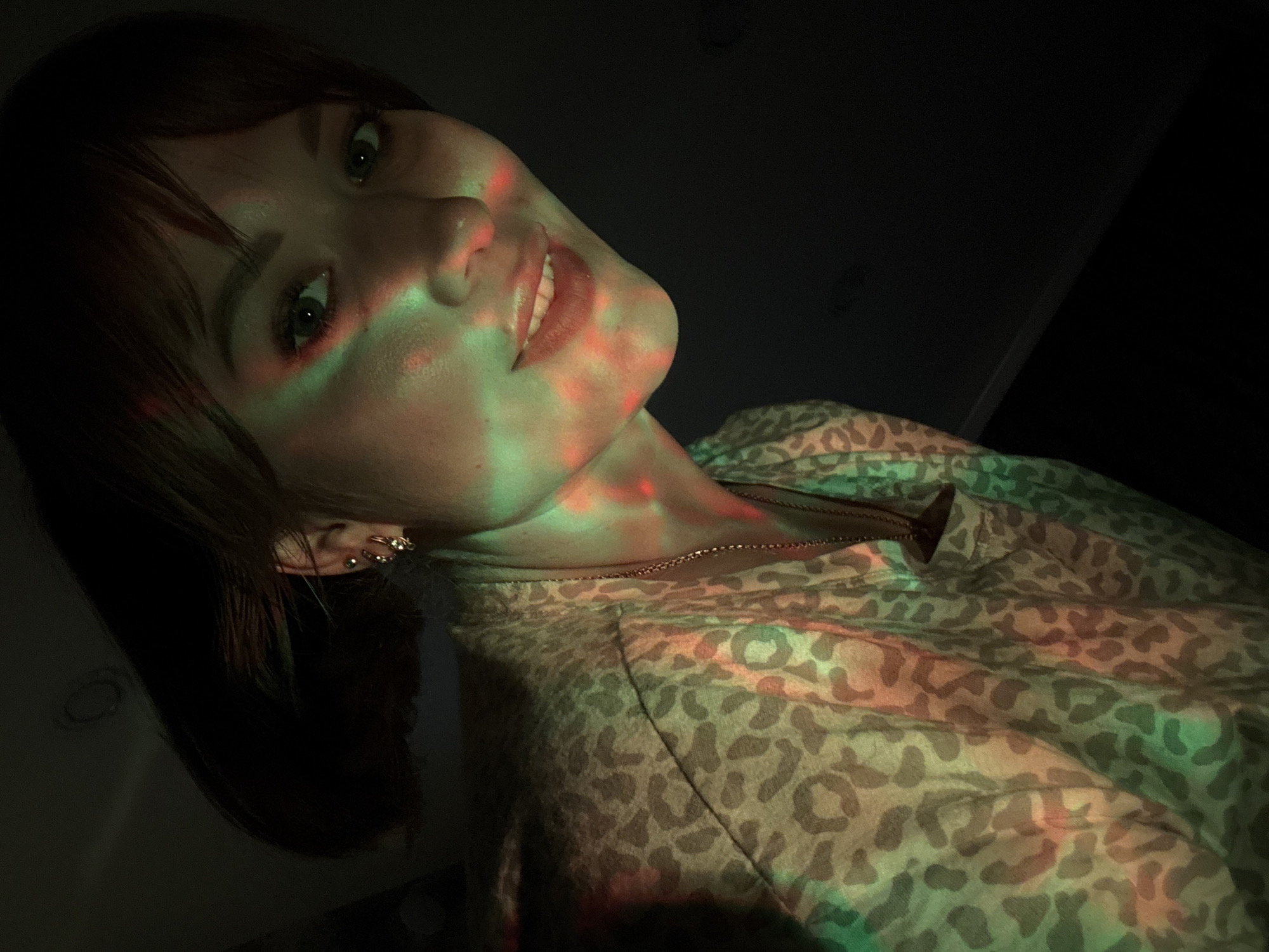The image depicts a young adult female from the chest up, positioned slightly below eye level and angled upwards. She has reddish-brown, shoulder-length hair with bangs covering her forehead. Her complexion appears fair, and she has visible eye makeup. Multiple small, gold-colored hoops and studs are present in her right earlobe and helix.
She is wearing a light-colored top with a dark, mottled leopard-print pattern. A thin, gold-colored chain necklace with a small, dark, pendant hangs around her neck. Her face and upper body are illuminated by projected red and green lights, creating distinct colored patterns and spots across her skin and clothing. She looks directly towards the viewer with a slight, closed-mouth smile.
The background is uniformly dark, providing minimal contextual details; however, faint shapes in the upper left suggest an indoor setting with minimal ambient light. There is no discernible visual evidence within the image to indicate its location within the city of Chaikovsky, Russia, or any specific geographical area.
