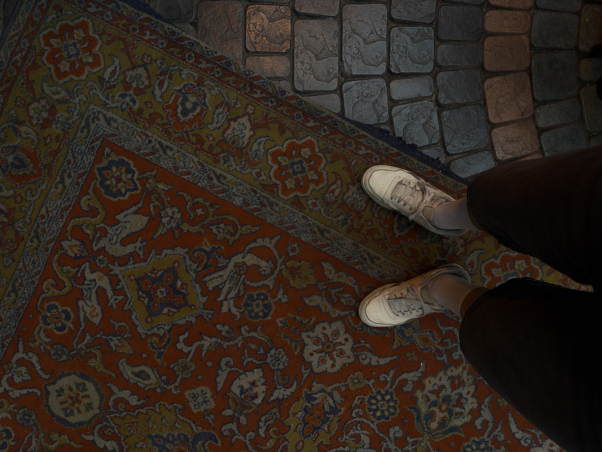 An overhead shot shows the lower portion of a person standing on a patterned rug with part of the rug on a tiled floor. The person is wearing black pants, white socks, and white sneakers. The rug is patterned with red, blue, orange, and yellow. The tiles are gray and textured. The setting is likely indoors, possibly a store or home. The location is unknown.