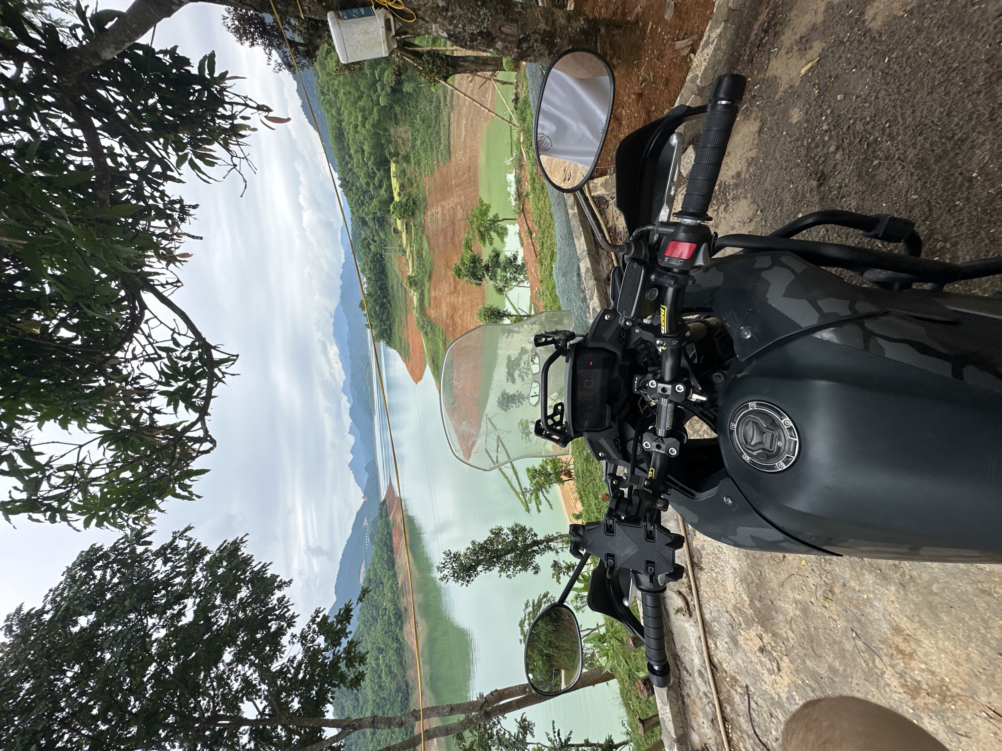 A dark motorcycle with a camouflage pattern on its tank is parked on a concrete surface, facing the viewer. The motorcycle has a clear windshield and handguards. In the background, a large body of water with a greenish hue stretches out, surrounded by rolling hills covered in lush green vegetation. The sky is overcast with a few scattered clouds. Several trees, including one with large leaves in the foreground, frame the scene. To the right of the motorcycle, a white plastic container is attached to a tree. The location is identified as Bản Phúc, Vietnam. The image appears to be a scenic view captured from the perspective of someone on or next to the parked motorcycle.