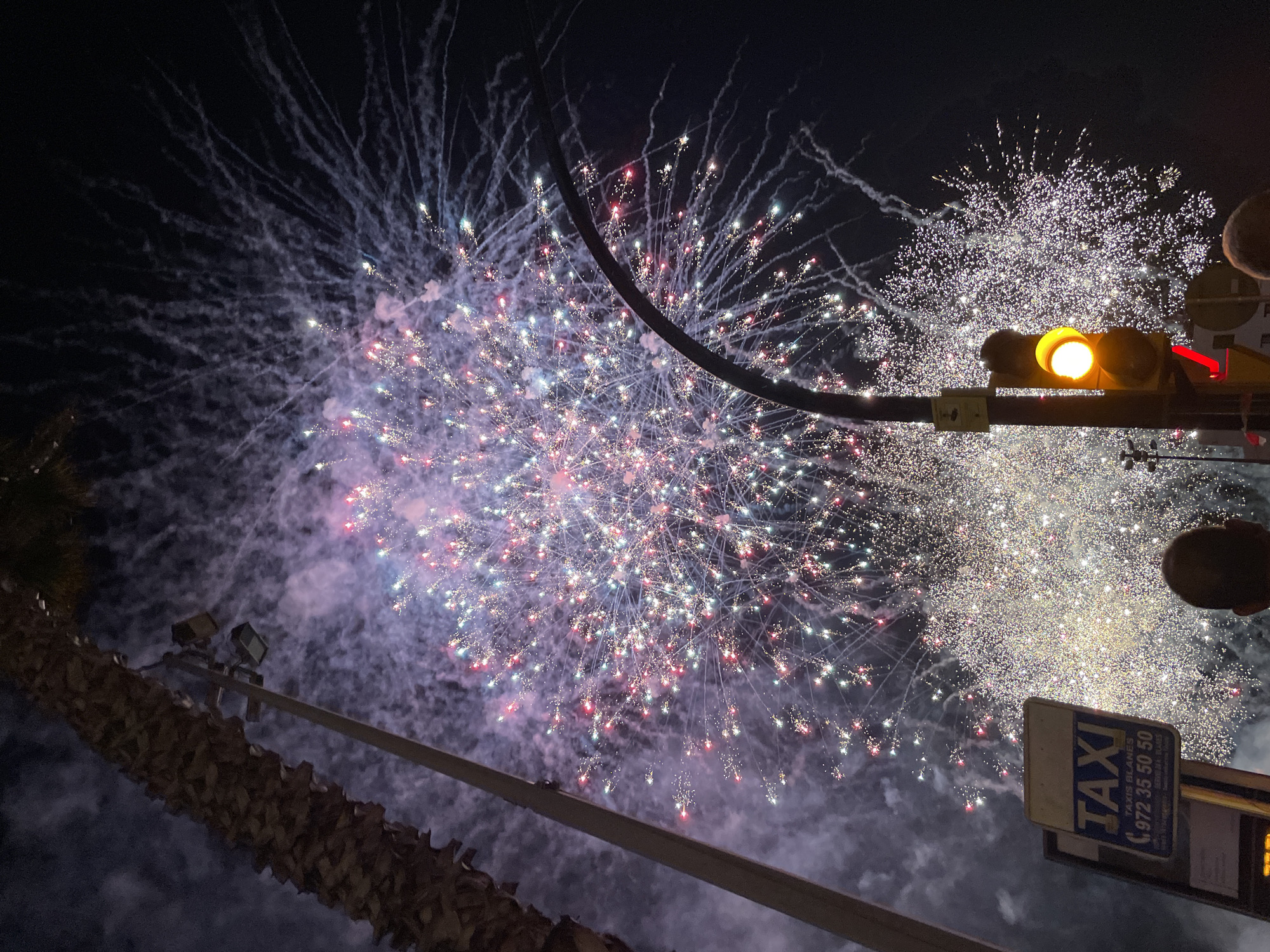 This is a night scene captured at the Blanes, Spain waterfront, likely during a fireworks display. A large burst of fireworks illuminates the dark sky with a dense cloud of white and pink smoke, dotted with numerous red, white, and blue sparks. In the foreground, a traffic light displays a yellow signal, and a taxi sign is visible, displaying "TAXI BLANES" and a phone number. A tall palm tree trunk is on the left side of the frame, and a street lamp is positioned near the center. The heads of two individuals are visible in the lower portion of the image, looking towards the fireworks. The scene suggests a public event with a festive atmosphere.