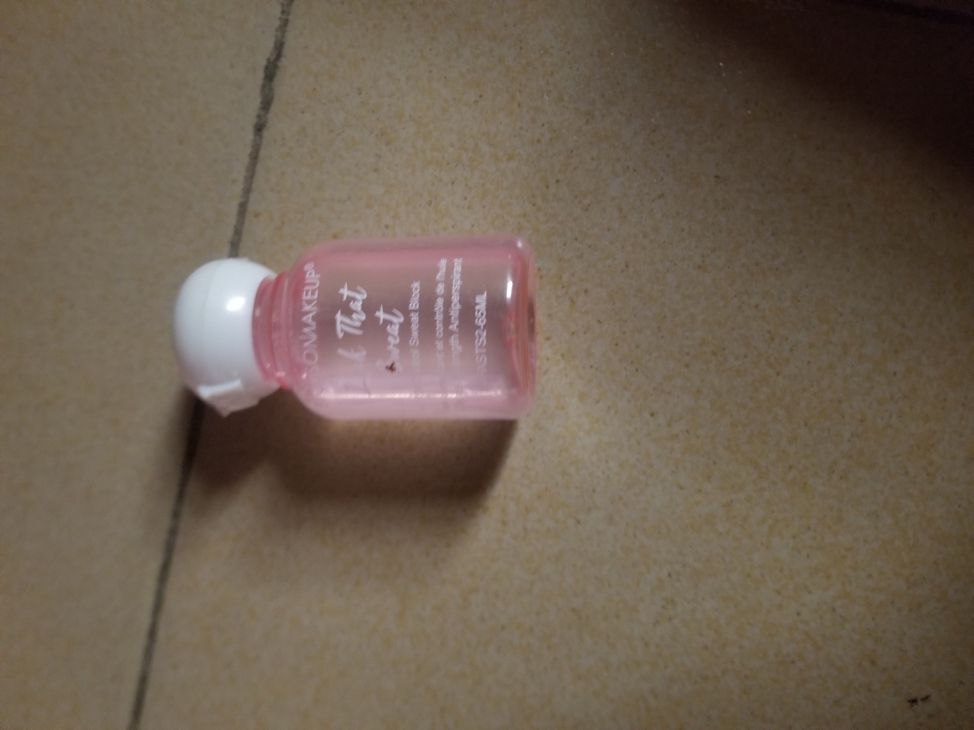 A close-up, eye-level, indoor shot captures a small, translucent pink bottle with a white cap, lying on its side on a tiled floor. The bottle is positioned horizontally, with its cap to the left and the base slightly raised. Text on the bottle is partially legible, indicating "ONMAKEUP" at the top, followed by "k That" and "wreat" in a script font. Below this, smaller text reads "rol Sweat Block" and "th Antiperspirant". The base of the bottle has the inscription "ASTS2-65ML". The tiled floor is a speckled beige and brown, with faint grout lines. The lighting is dim, casting a soft shadow to the right of the bottle. The overall impression is of a product placed casually on a domestic surface. There is no indication of Jauro Yinu, Nigeria in the image.
