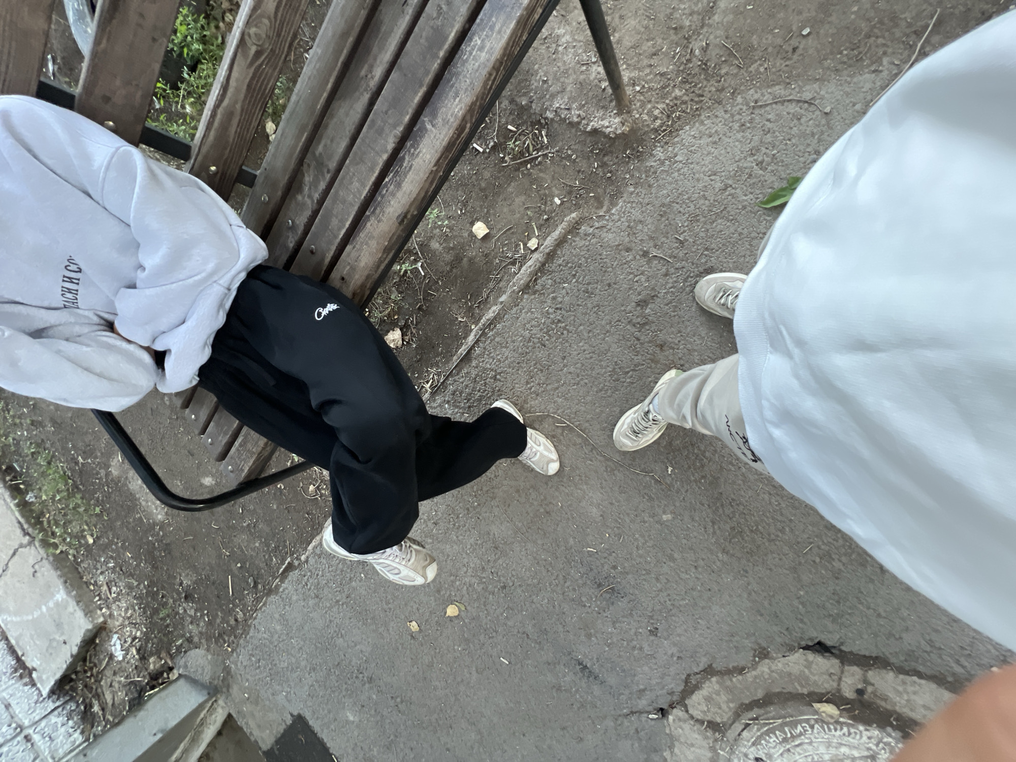 Two individuals are positioned outdoors. The first individual, seated on a wooden bench, is wearing a light-colored hooded sweatshirt with dark text across the chest and black sweatpants with a white logo on the left thigh. They are wearing white athletic shoes and have their arms crossed. The second individual is standing, facing away from the camera, and wearing light-colored pants and white athletic shoes. They are captured from the knees down. The environment appears to be a public area with a concrete sidewalk and dirt ground. The bench has a metal frame. The overall scene suggests a casual outdoor setting. There are no visible landmarks or specific indicators that place the scene within the city administration of Qarağandı, Kazakhstan.