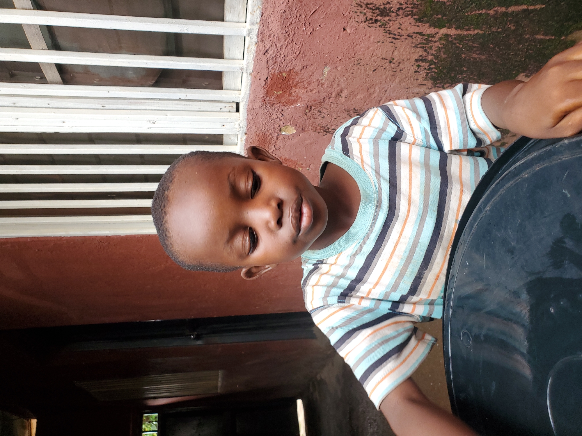 The image shows a close-up, vertically oriented shot of a young Black child sitting next to a large, black object, possibly a plastic container or tub. The child is wearing a striped t-shirt with horizontal stripes in white, light blue, orange, and dark blue. Their head is tilted, and their eyes are partially closed, giving an impression of restfulness or contemplation. The child's skin tone is dark.
Behind the child, there is a textured, reddish-brown wall. To the left of the wall, there is a white structure that appears to be a window with horizontal slats. The background also shows some darker areas, suggesting an outdoor or semi-outdoor setting. The ground surface in the foreground is a light, dusty color.
The lighting suggests it could be daytime, but the exact time is not definitively indicated. There are no discernible weather cues. The setting appears to be a residential area in Jos, Nigeria, given the architectural style of the window and the wall texture. The overall mood of the image is calm and intimate, focusing on the child's presence. There is no visible text in the image.