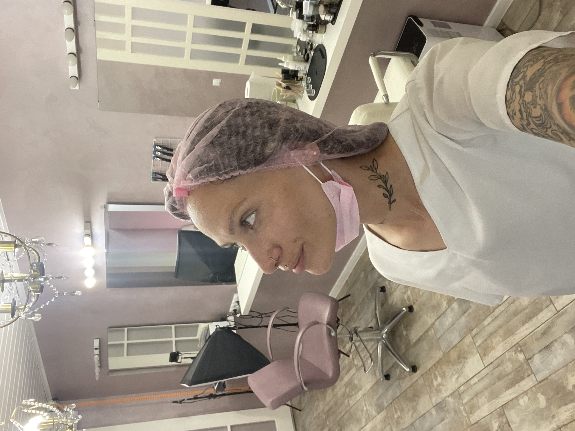 A medium shot captures a person wearing a pink disposable hairnet and a light pink face mask, positioned in what appears to be a salon setting. The individual is wearing a white garment and displays a visible tattoo of a plant on their neck. Their left arm, covered in tattoos, is also visible. The background includes salon furniture, such as pink chairs and a black lamp, along with cosmetic products arranged on shelves. The floor is composed of light brown, wood-patterned tiles. A chandelier can be seen in the upper left corner of the image. The image is not clear enough to identify the specific location in Izhevsk, Russia.