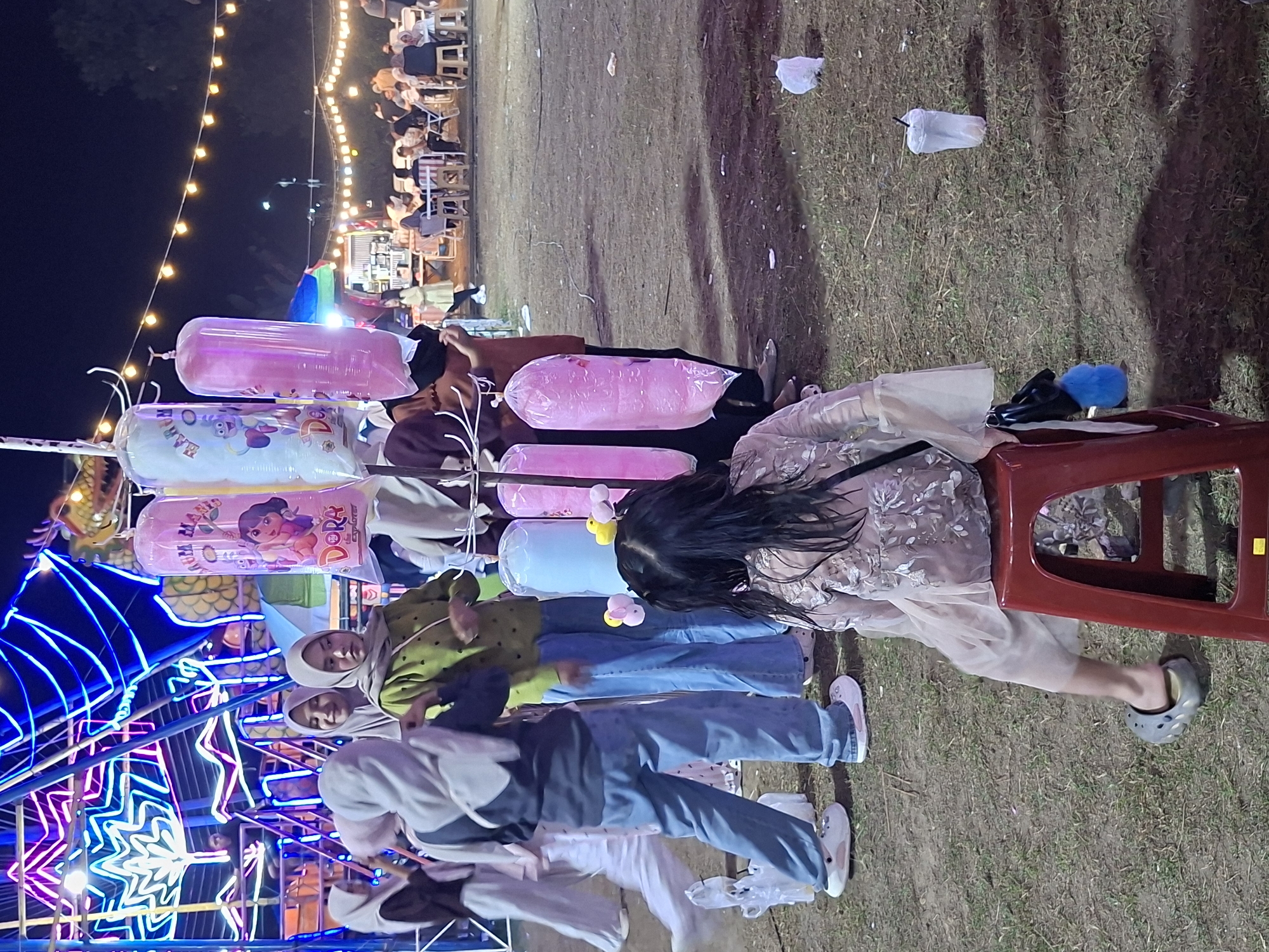 At night in Geulumpangsulu Timu, Indonesia, a fairground scene is depicted. A line of people, including several women wearing hijabs, stand near a vendor selling inflated, plastic-wrapped toys, some featuring cartoon characters like Dora. Blue and white neon lights illuminate a nearby structure. A woman sits on a red plastic chair. The ground is covered with grass and scattered plastic waste. Background lights indicate other people and activities in the distance.