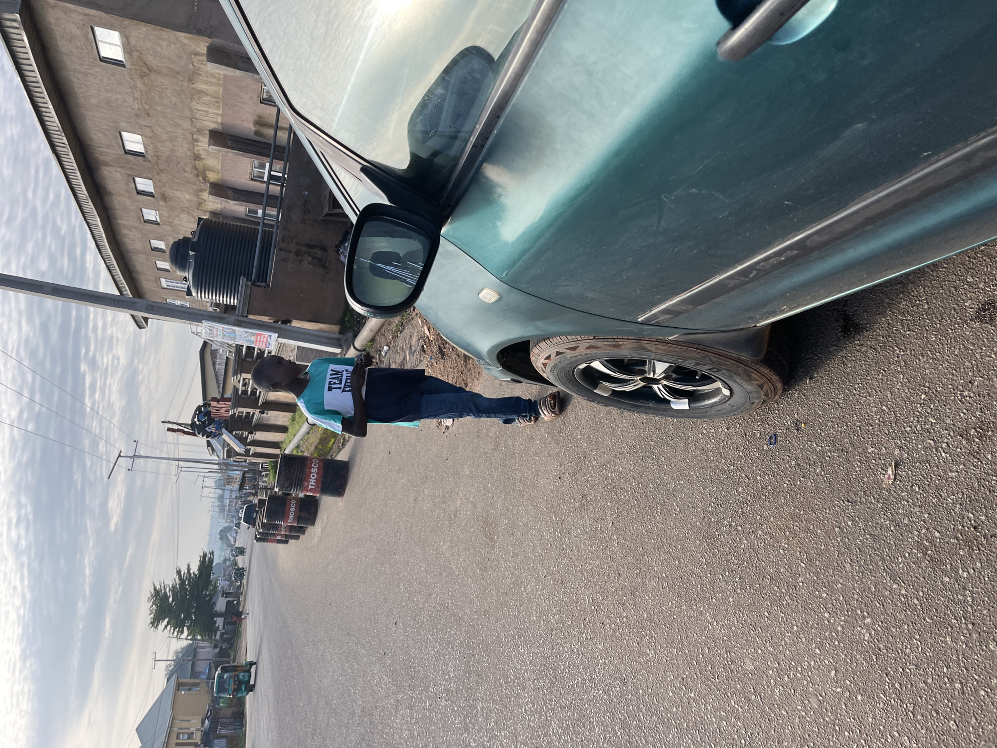 The image depicts a roadside scene in Jos, Nigeria, likely in the early morning or late afternoon due to the lighting. In the foreground, a dark green car is partially visible, its side and wheel taking up a significant portion of the right side of the frame. A young Black male, appearing to be a teenager, stands on the left side of the road, facing away from the camera. He is wearing a t-shirt with a graphic that includes the words "TEAM" and "EVERY." He also wears blue jeans and sandals.
Behind the boy, a row of black barrels, some with red markings and text that appears to be "THOSCO," are lined up along the roadside. The road itself is paved and appears to be a moderately trafficked street, with other vehicles, including a tricycle (Keke Napep), visible in the distance. Several buildings line the left side of the road, with one prominent building on the right featuring a large black water tank on its roof and multiple windows. The sky is partly cloudy with a soft, diffused light. The overall environment suggests a commercial or residential area bordering a main road.