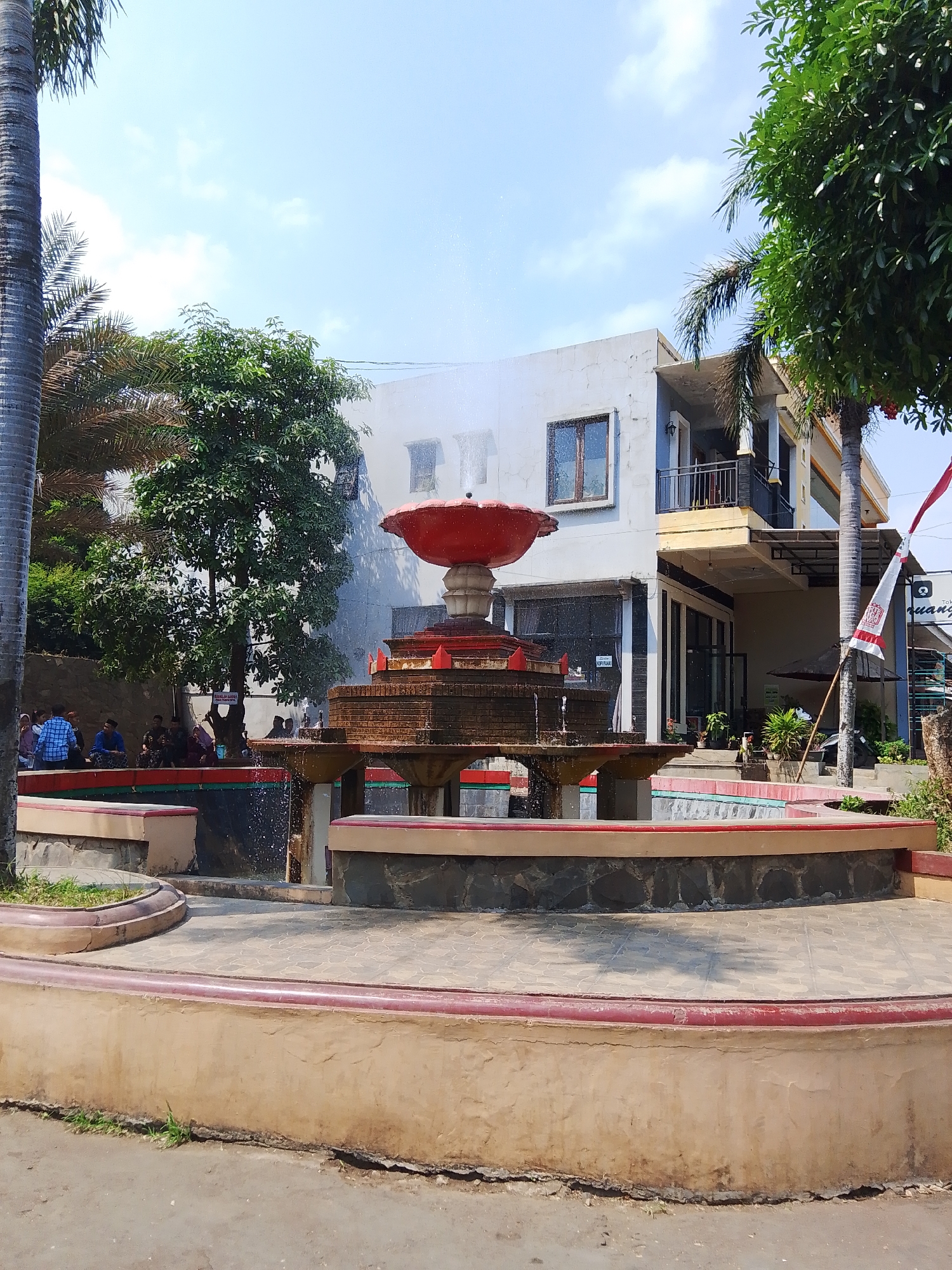 The image shows an outdoor scene with a fountain as the central element. The fountain is multi-tiered with a red bowl-shaped top. It is surrounded by a decorative, multi-level basin made of stone and concrete, with red and cream accents. Water is visibly spraying from the fountain. Behind the fountain, there is a light-colored, multi-story building with windows and balconies. Trees and palm trees are present to the left and right of the fountain, with lush green foliage. In the background, to the left of the fountain, a group of people are seated and standing, some appearing to be socializing. A red and white flag is visible on a pole to the right of the fountain. The ground in the foreground is paved with textured tiles and concrete. The sky is blue with scattered clouds, indicating a daytime scene. The overall impression is of a public space or garden area within a town or city, likely a recreational spot. Based on the provided information, this scene is located in Mantingan, Indonesia.