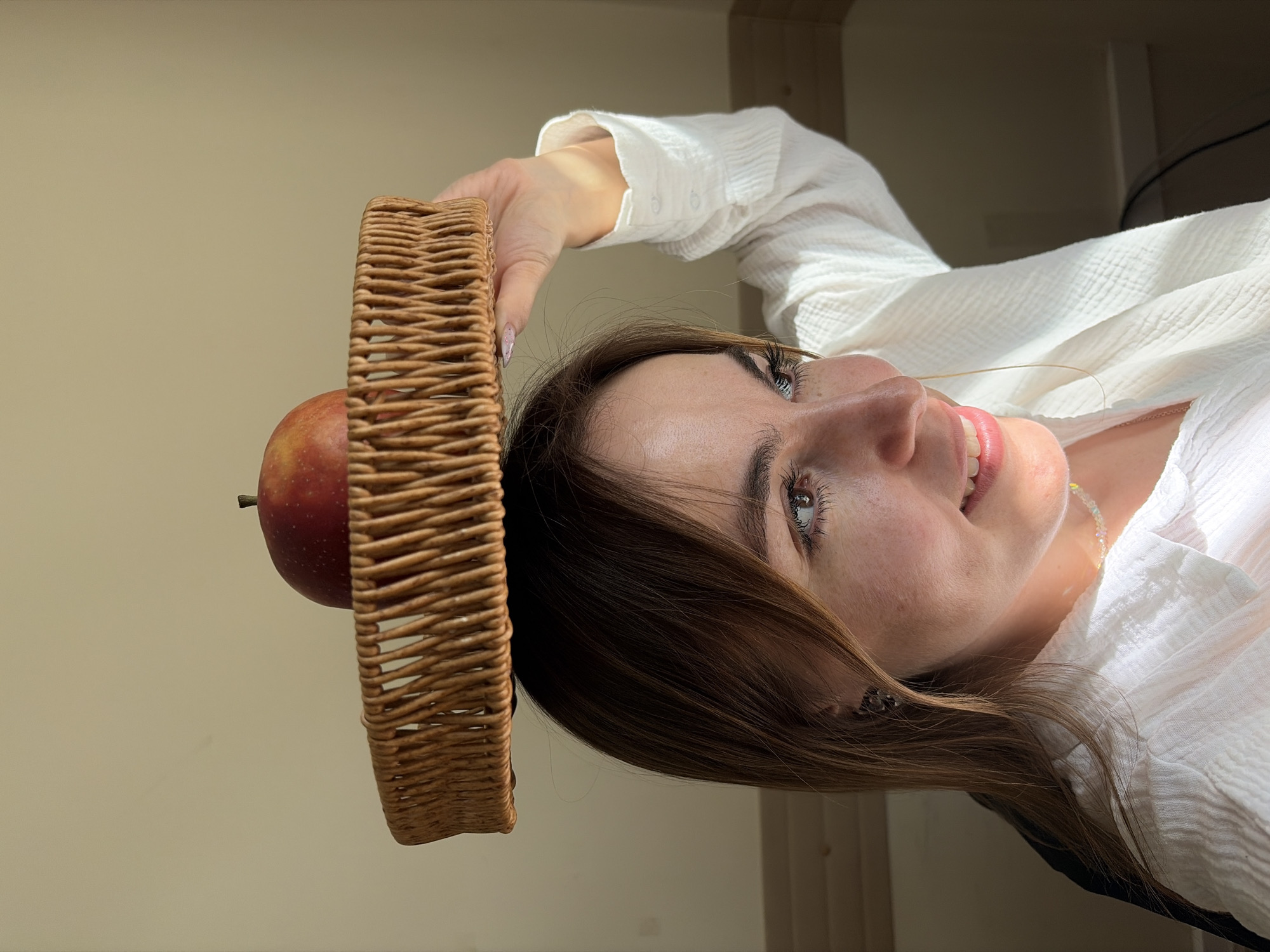 A woman in a white, long-sleeved shirt is positioned with her head near the top of the image. A round, woven basket sits on the top of her head, and a red apple is positioned within the basket, visible on the left. The background is a neutral tan color. The image appears to be taken indoors. There is no information that indicates the location to be Izhevsk, Russia.