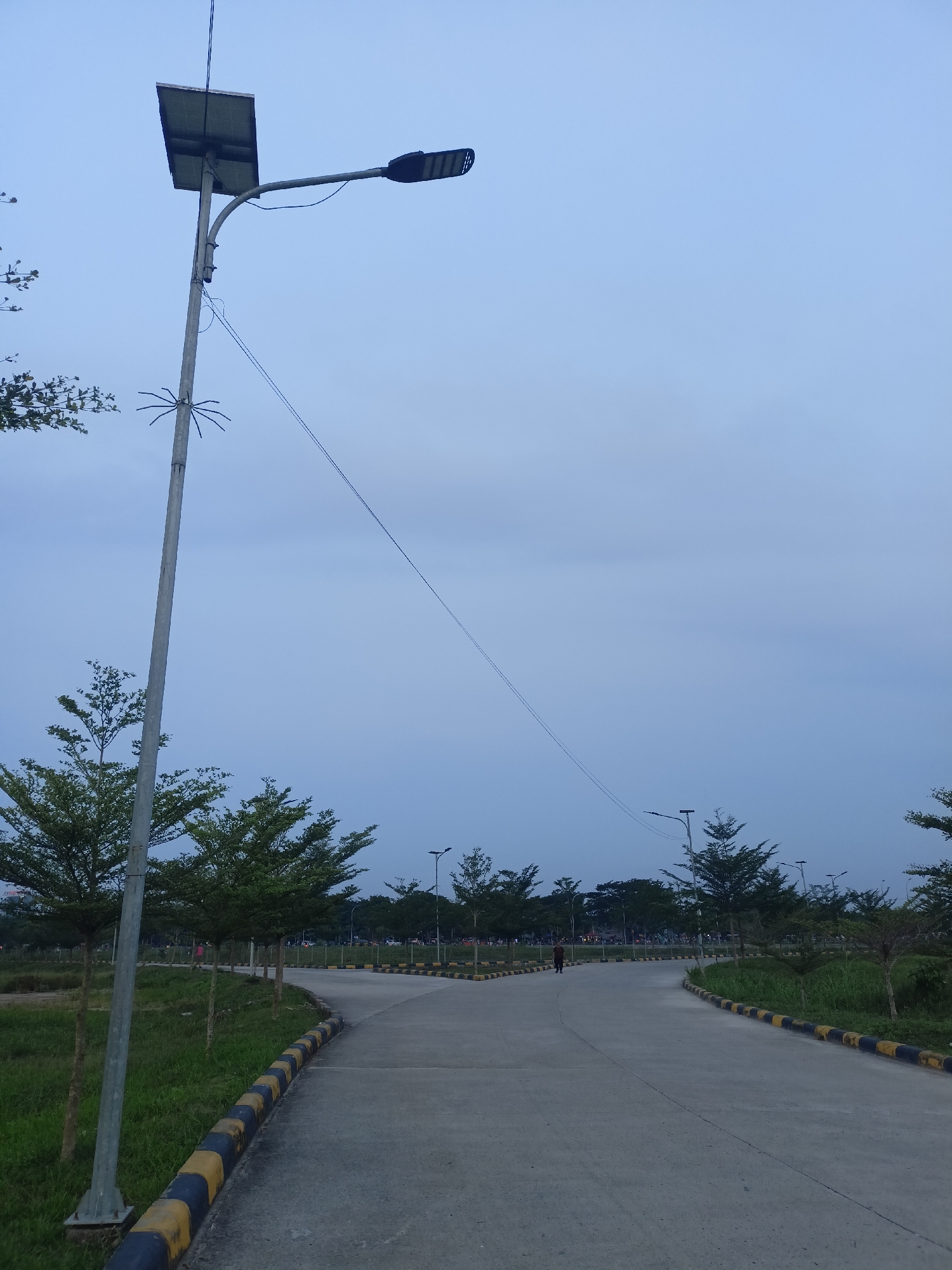 The image shows a paved pathway in what appears to be a park or public space in the city of Sena, Indonesia. The pathway forks, with one side curving to the left and the other to the right. The ground is concrete and bordered by grass and a yellow and black striped curb. Several streetlights, some with solar panels, are visible along the pathway. Trees with sparse foliage are planted on either side of the path. In the distance, a lone person is walking on the pathway. The sky is overcast and a pale blue, indicating it is either early morning or late afternoon. The scene is tranquil and devoid of any immediate activity.