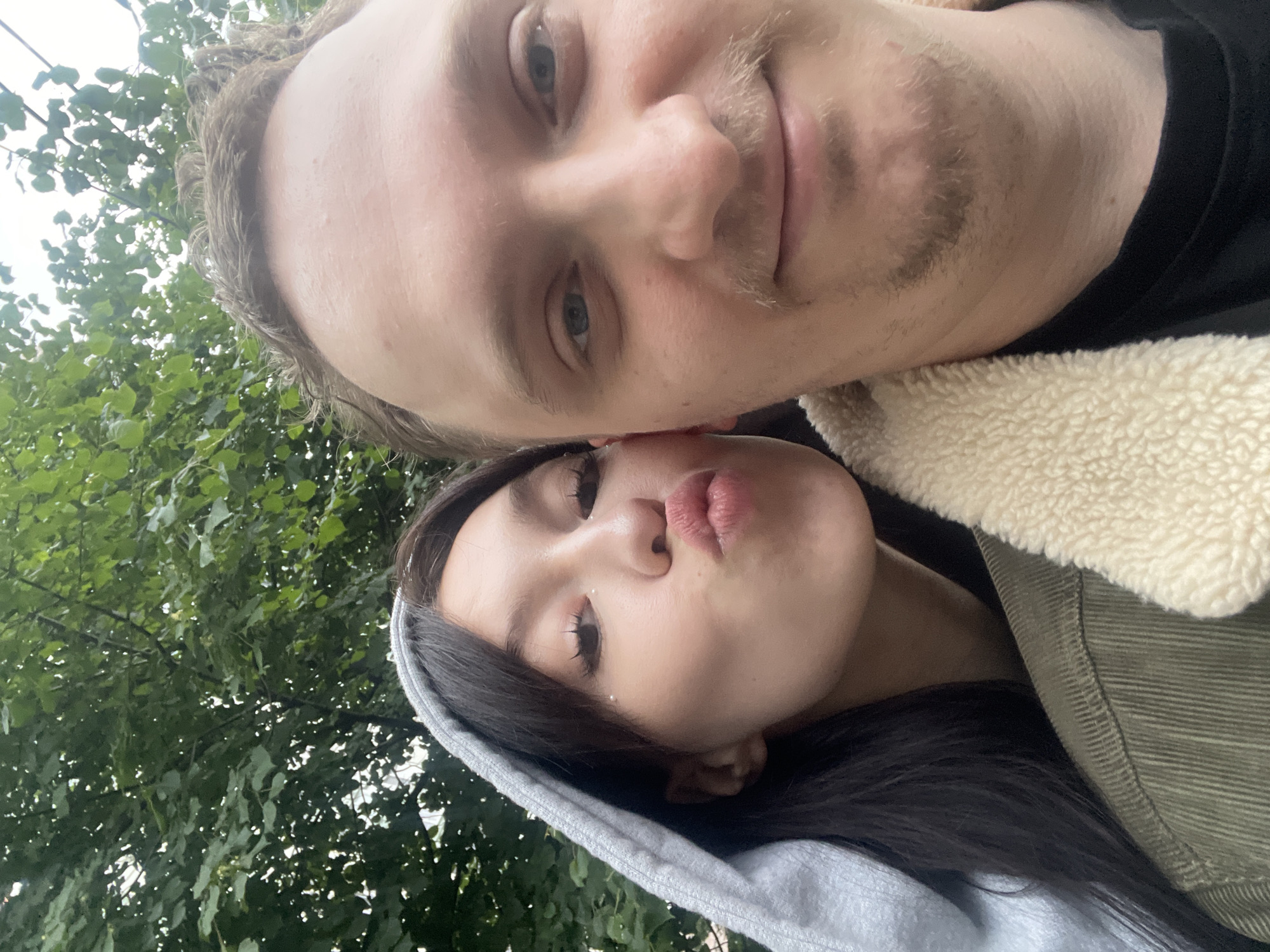 In a close-up selfie taken outdoors, two individuals are depicted against a backdrop of lush green foliage, suggesting a park or wooded area. The person on the left, a woman with dark hair, is wearing a grey hooded sweatshirt and is leaning in, puckering her lips towards the camera. She has subtle makeup, including what appears to be glitter on her eyelids. The person on the right, a man with light brown hair and a mustache, is facing the camera directly, smiling slightly. He is wearing a green corduroy jacket with a cream-colored sherpa lining. The lighting suggests it is daytime, possibly overcast. The scene is devoid of any specific urban landmarks, making it difficult to pinpoint a location within Khimki, Russia.