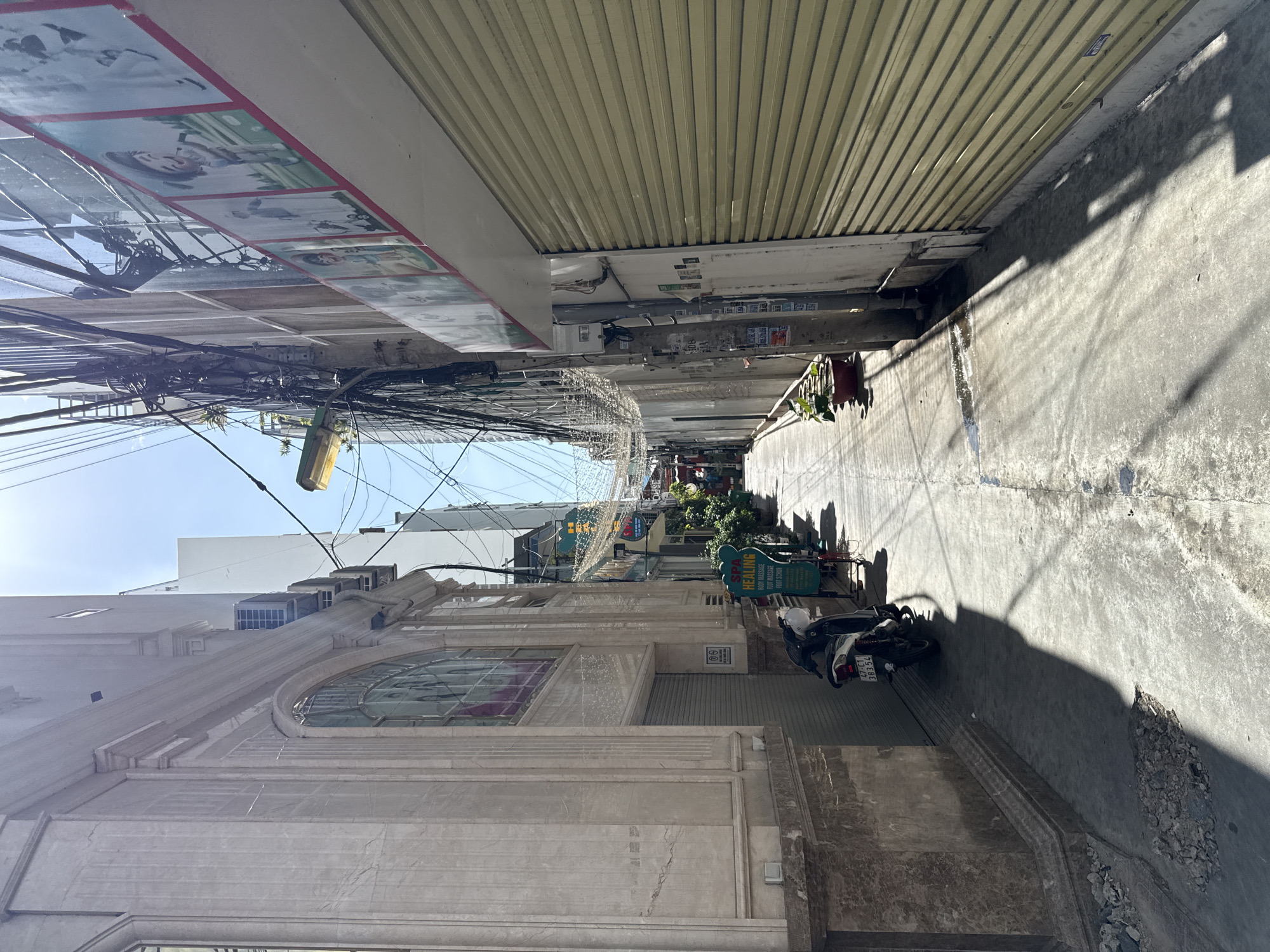 The image depicts a narrow street in Nha Trang, Vietnam, lined with buildings on both sides. The street is paved with concrete and shows signs of wear, with some debris scattered on the ground. The buildings are a mix of architectural styles, with one on the left featuring a marble facade and decorative window. The building on the right has a yellow roll-up door and is adorned with a series of framed pictures along its upper edge. Overhead, a dense network of electrical wires crisscrosses the sky, and a yellow street lamp is visible.
In the foreground, a black motorcycle is parked on the left side of the street, casting a long shadow. A green sign for "SPA HEALING" is positioned next to the motorcycle, advertising body massage and foot scrub services. Further down the street, plants in pots are visible, and the lane narrows and curves out of view. The sunlight casts strong shadows, indicating it is likely midday. The overall impression is one of a bustling urban environment, characteristic of many streets in Vietnamese cities.