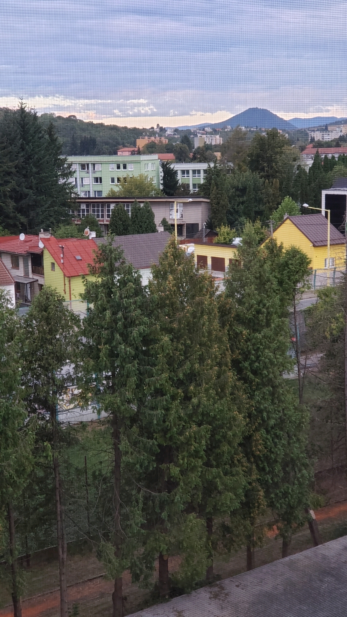 The image depicts an urban landscape with buildings and trees under a cloudy sky. In the foreground, several evergreen trees are prominent. Behind them, a cluster of houses with red and yellow roofs are visible. Further in the background, larger buildings, including a light green structure with multiple windows, are situated. In the distance, a city skyline with more buildings and a large, conical hill or mountain can be seen. The scene appears to be a residential or mixed-use area within a city. The overall lighting suggests it could be late afternoon or early morning. The image quality is somewhat grainy, possibly due to a screen or filter.
The visual elements in the image are consistent with the city of Prešov, Slovakia, specifically the area of Sídlisko III, a large housing estate. The distinctive green apartment building in the middle ground is characteristic of this neighborhood. The conical hill in the distance is likely the Šariš Castle hill, a prominent landmark visible from many parts of Prešov. The presence of residential buildings, apartment blocks, and the surrounding natural landscape with hills aligns with the topography and urban development of Prešov.