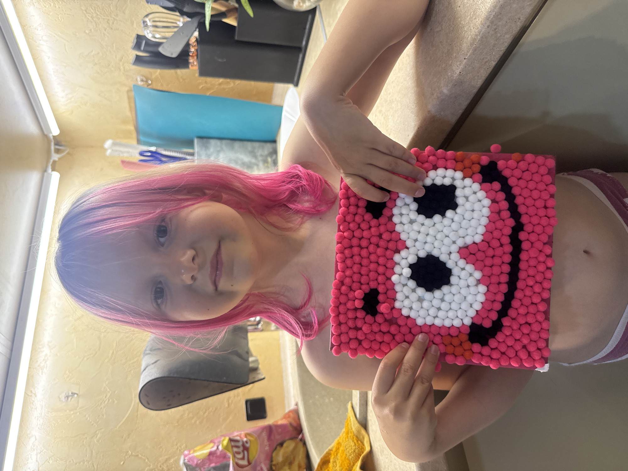 A girl with pink hair is holding up a square object covered in small pink, white, and black pom-poms that form a face with large eyes. The girl is positioned in front of a light-colored wall, and kitchen countertop and kitchen utensils are visible in the background. A bag of Lay's potato chips is visible in the bottom left corner. The location is Unknown, Unknown.