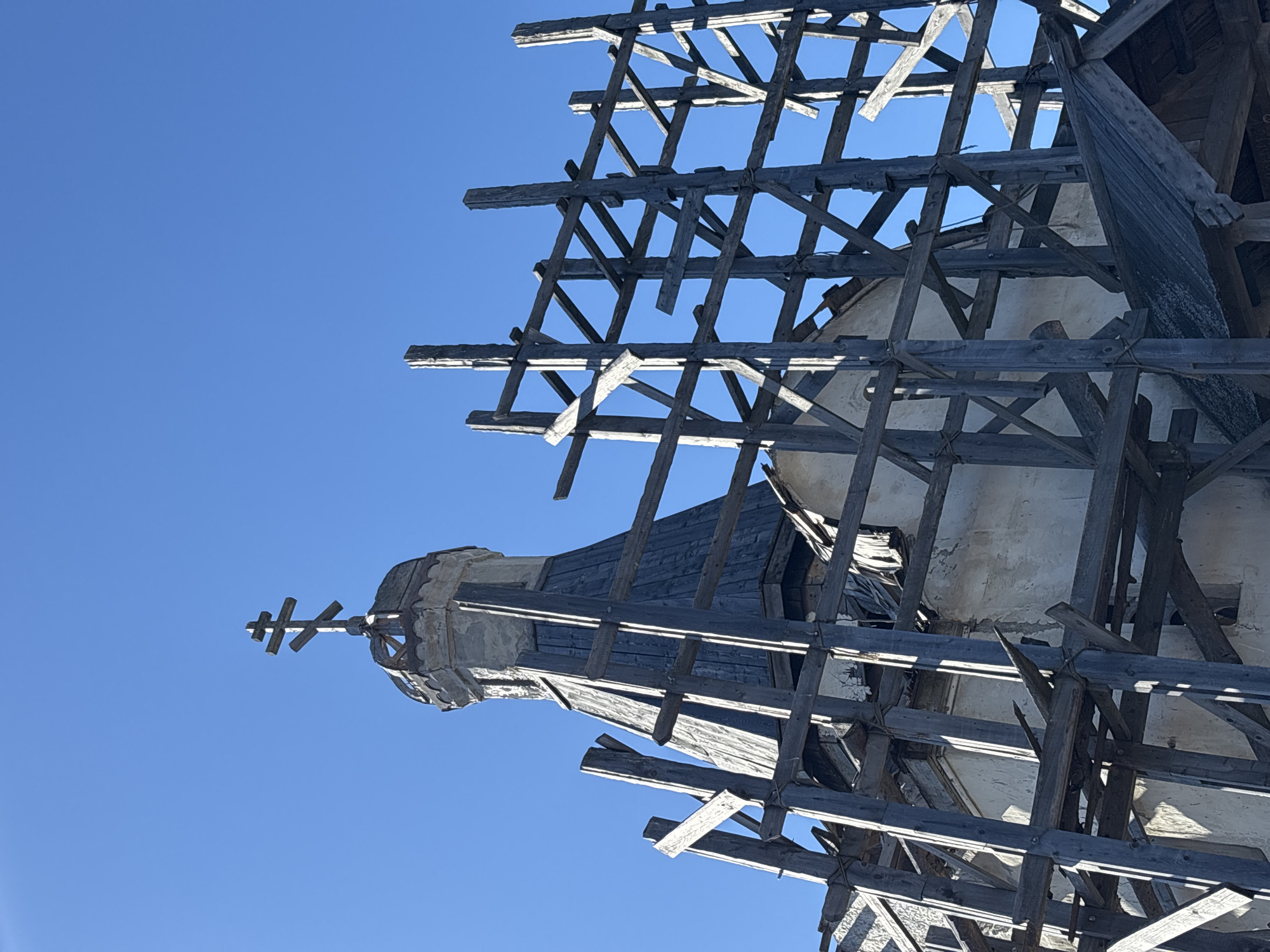 The image shows a dilapidated church steeple under repair in Gubakha, Russia, viewed from below against a clear blue sky. The steeple features a prominent Orthodox cross at its apex. The steeple itself appears aged and worn, with visible damage and deterioration. Extensive wooden scaffolding surrounds the structure, indicating ongoing restoration work. The scaffolding is a complex network of beams and supports, partially obscuring the steeple.