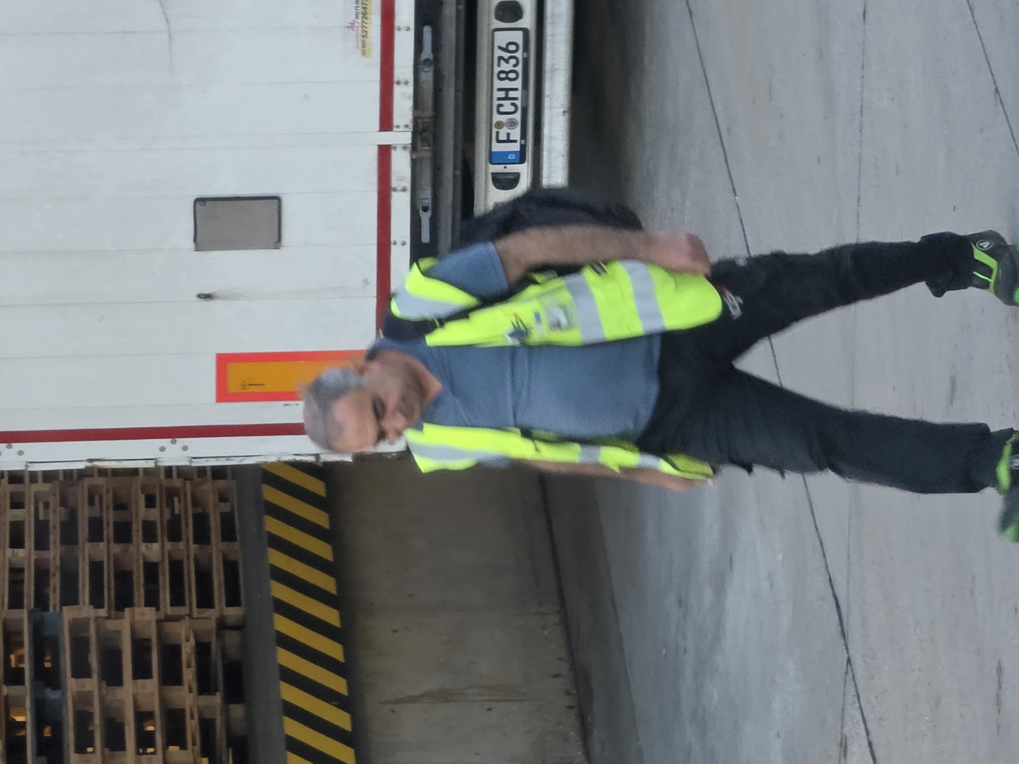 A man wearing a blue shirt, a reflective yellow vest, and black pants walks on a concrete surface. He has a backpack over his right shoulder. In the background, there is a white truck with a license plate that reads "F CH 836". To the left of the truck, there is a stack of wooden pallets and a concrete wall with a yellow and black striped warning barrier. The scene appears to be an industrial or logistics area, possibly a loading dock, in Frankfurt, Germany.