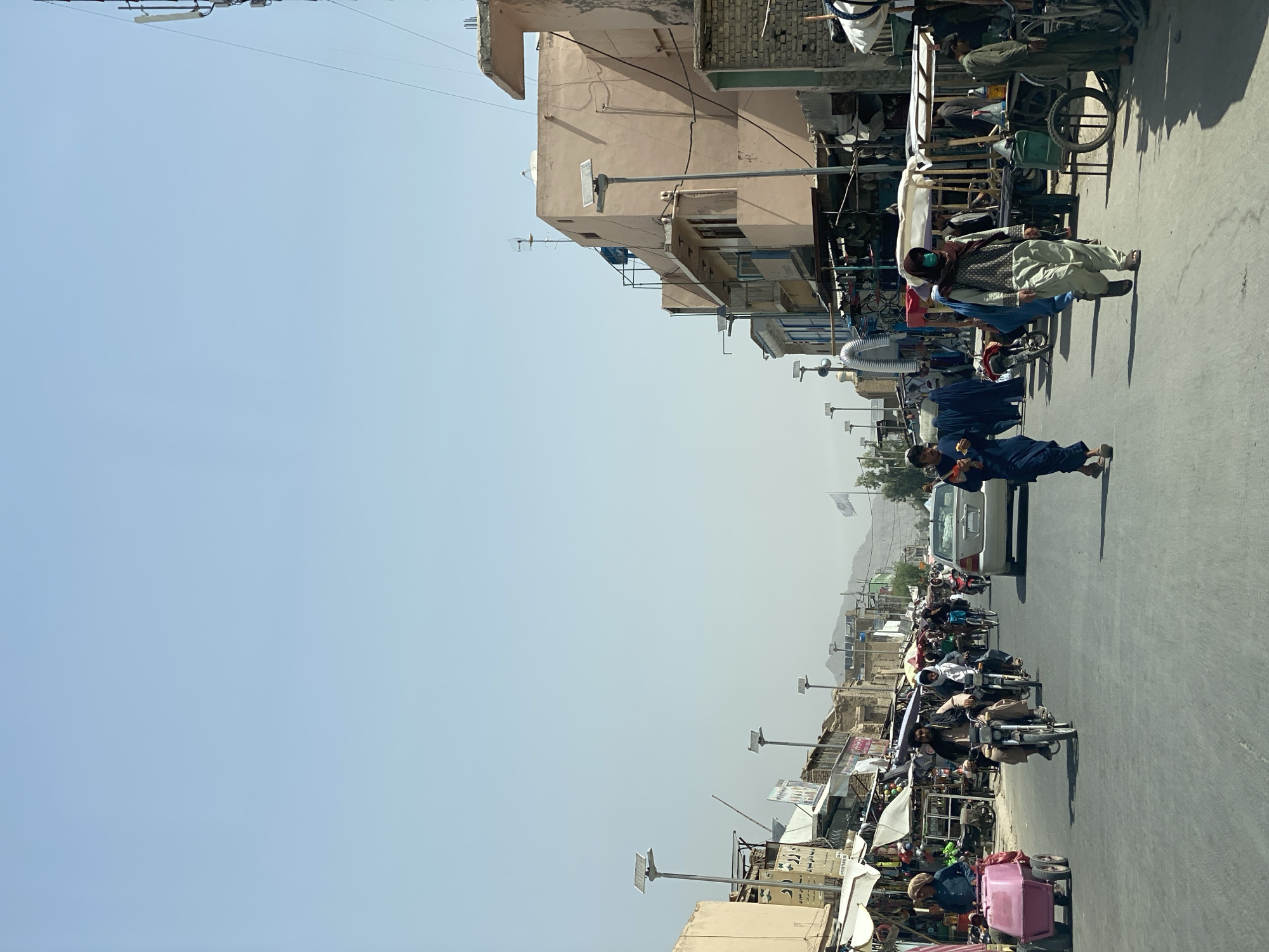 The image depicts a street scene in Kandahar, Afghanistan. The street is lined with market stalls and buildings. Several people are present, some walking and others riding motorcycles. A white sedan is also visible on the road. The sky is clear and blue. The overall impression is that of a busy, everyday market day.