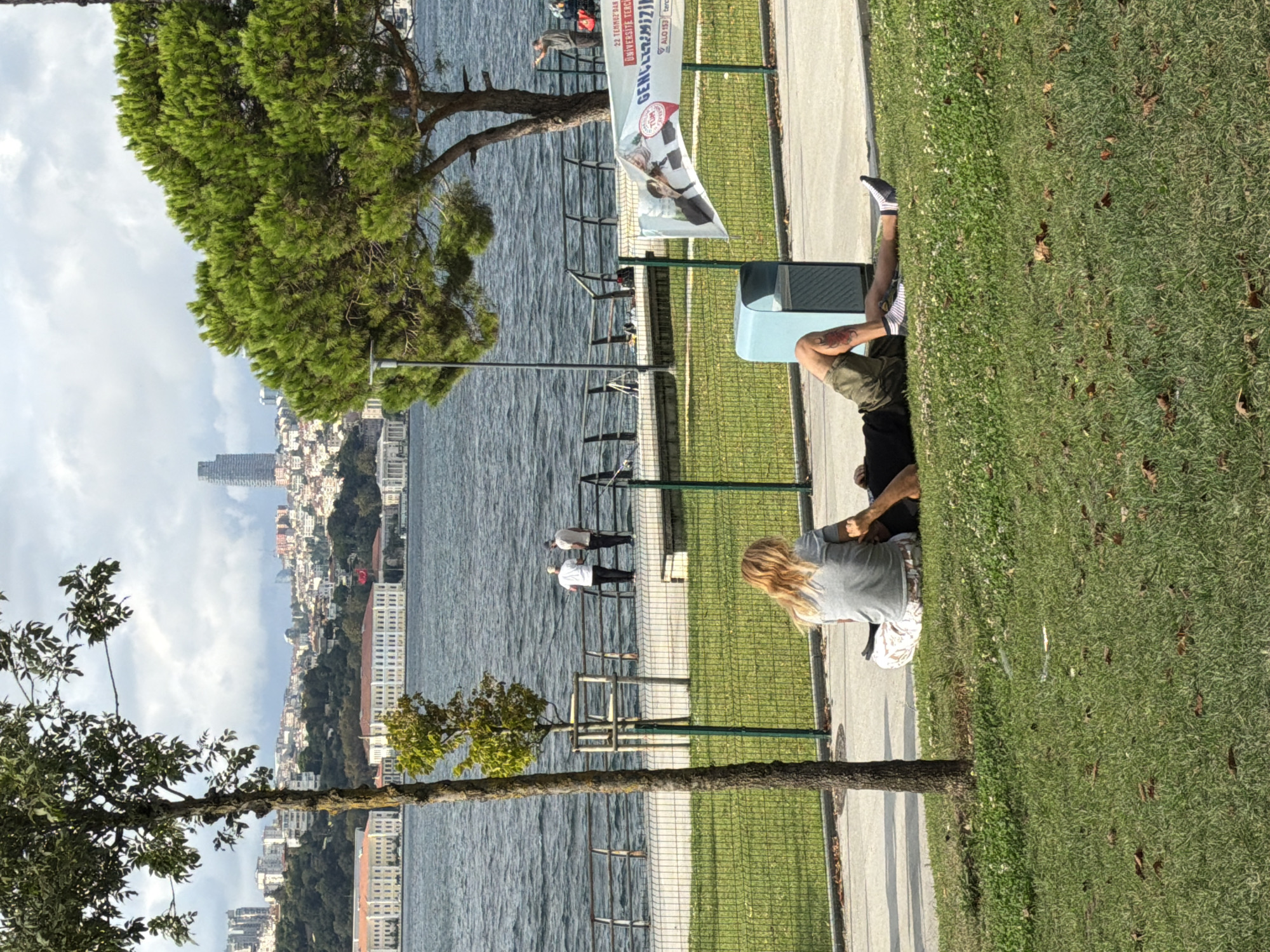 The image depicts a sunny day in Üsküdar, Turkey, along the Bosphorus Strait. In the foreground, two individuals are relaxing on a grassy area adjacent to a paved walkway. One person, facing away from the camera with blonde hair, is sitting with their legs crossed and wearing a grey t-shirt and patterned pants. Beside them, another person, lying down with their head towards the left, wears a black t-shirt and olive-green shorts, revealing a tattoo on their left thigh. A light blue trash receptacle is situated behind the seated individual.
Further back, a metal railing separates the park area from the water. Two individuals in white shirts and dark pants stand near the railing, facing the water. The Bosphorus Strait stretches across the midground, with the city skyline of Istanbul visible on the opposite shore. Prominent buildings include a tall skyscraper and numerous residential structures, interspersed with trees and foliage. A large building with white facades and red-tiled roofs is visible closer to the water's edge, and a Turkish flag flies from its vicinity. On the right side of the image, a large evergreen tree with dense needles provides shade, and a banner displaying images of people and Turkish text is partially visible, attached to a fence. A street lamp stands near the tree. The sky is partly cloudy, with a mix of sun and scattered clouds.