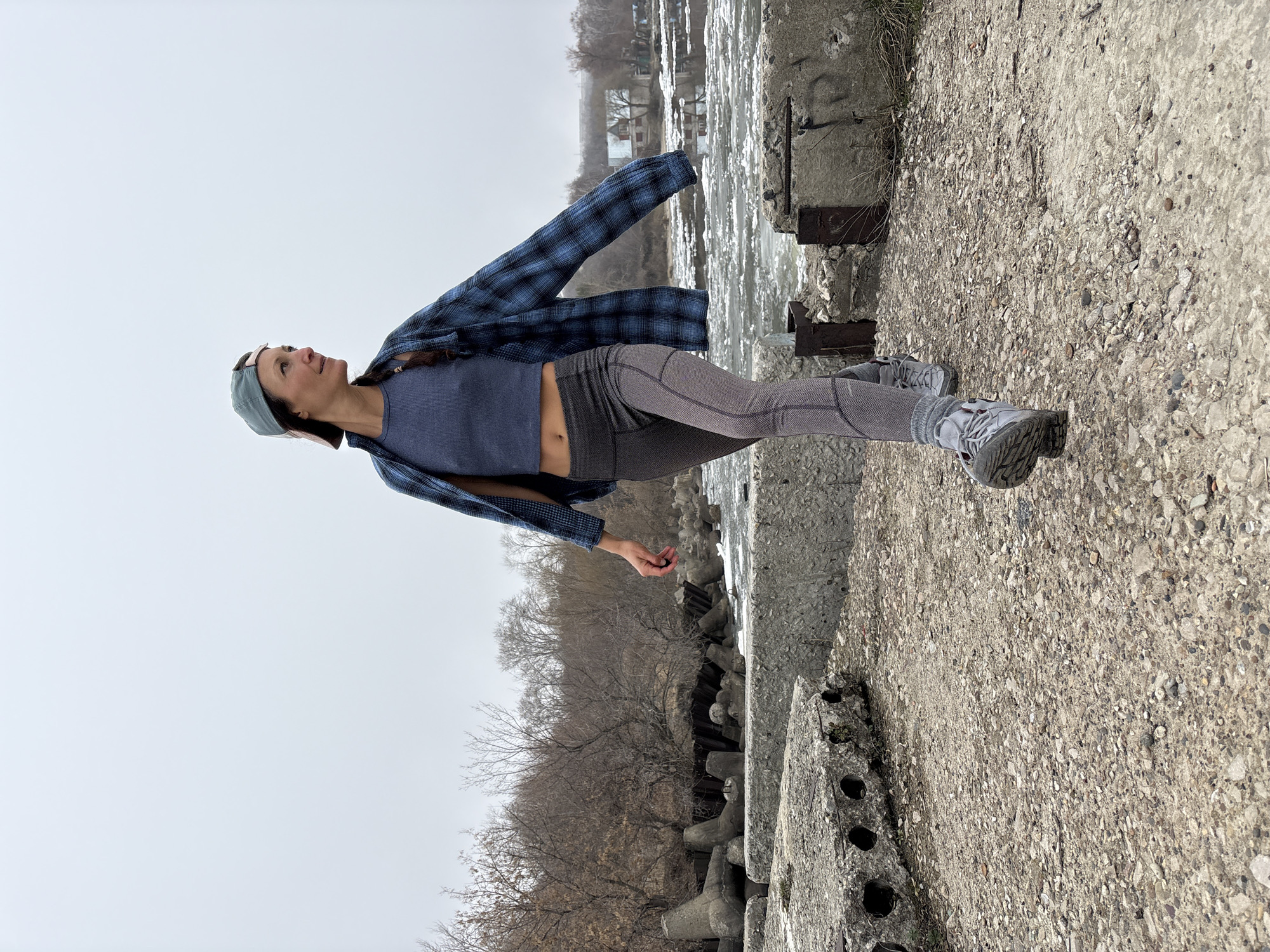 A full-length photograph captures a woman standing on a concrete surface in what appears to be an outdoor environment. The woman is wearing a navy blue tank top, grey leggings, a blue plaid flannel shirt left unbuttoned, grey sneakers, and a baseball cap. She is positioned facing upward, with her gaze directed towards the sky. In the background, there's a river or large body of water with structures on the opposite bank. The sky is overcast, suggesting a cloudy day. The concrete surface she's standing on is adjacent to an area of concrete blocks, and beyond that, trees without leaves. There is no evidence in the photograph that would confirm this is taken at any particular city.