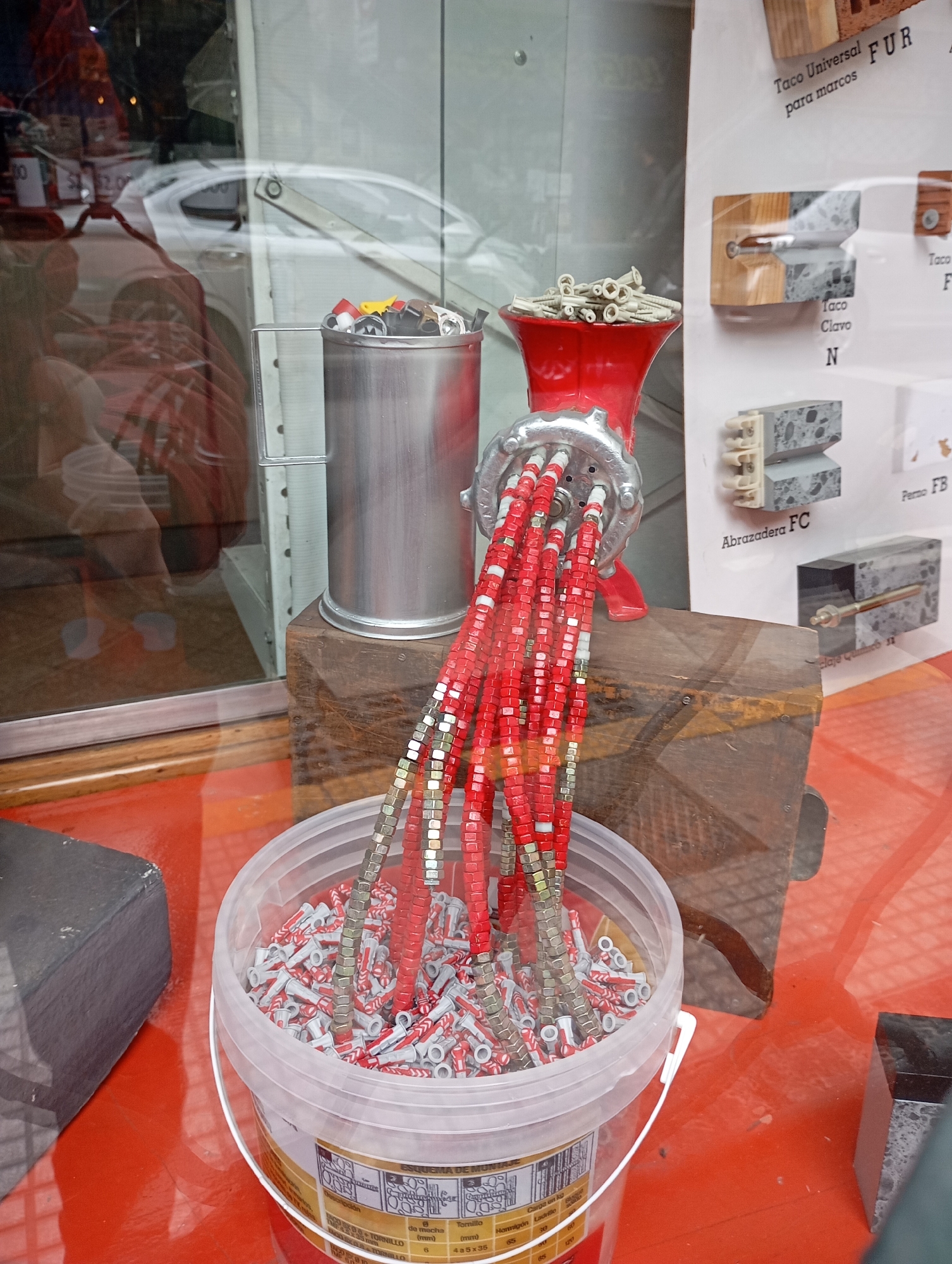 This image shows a display of construction hardware, possibly in a shop window in Buenos Aires, Argentina. The central focus is a repurposed red meat grinder from which numerous strands of red plastic wall anchors and silver metal nuts hang down. These strands are suspended above a clear plastic bucket overflowing with more of the same wall anchors. Behind the meat grinder is a silver metal can containing a variety of colorful caps or lids. The entire arrangement rests on a dark wooden block, which in turn sits on a red surface. Reflections on the glass surface suggest an urban environment, with glimpses of a white car and a person in the background. To the right of the display, a poster or sign showcases different types of wall anchors and their applications, with text in Spanish, including "Taco Universal FUR," "Taco Clavo N," and "Abrazadera FC." The overall scene appears to be an artistic or creative display of common building materials.