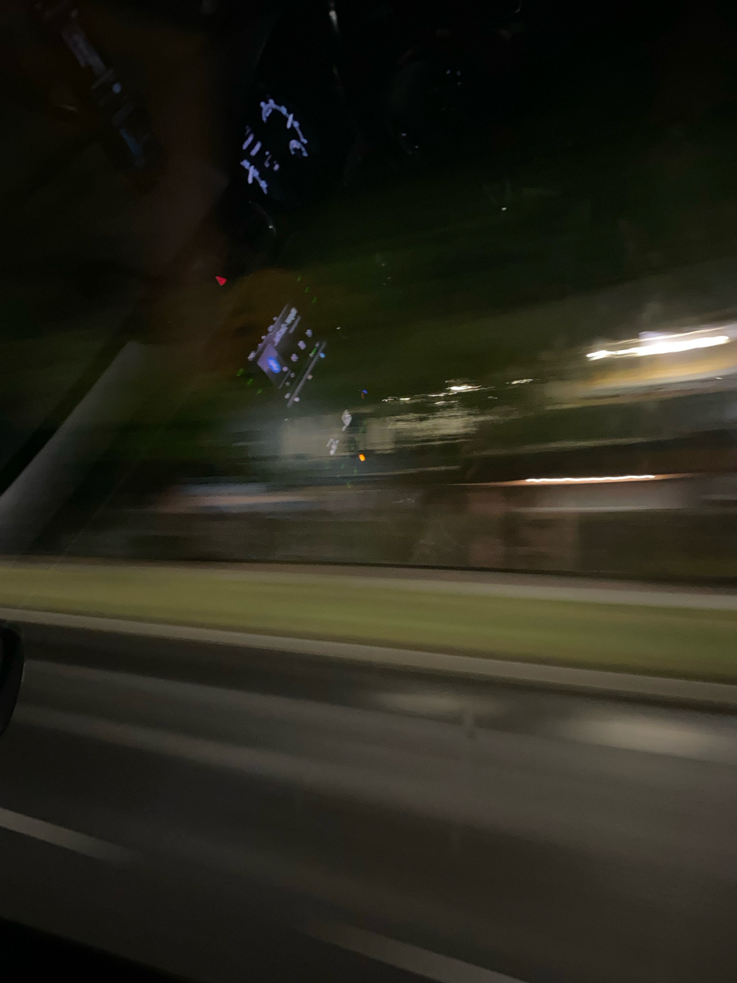 The image captures a blurred nighttime scene from inside a moving vehicle, likely in Katowice, Poland. The perspective is from the passenger seat, looking out the window. The foreground shows the interior of the car, with a partial view of the dashboard and a digital display. The dominant feature is the streaking motion blur of the outside environment.
The exterior view reveals streetlights and building lights creating horizontal streaks of light. A strip of green, possibly grass or a median, runs along the side of the road, also blurred by motion. The road surface is visible as a dark, grey expanse with lighter lines, further distorted by the movement. The overall impression is one of speed and transient observation of the urban landscape at night. No specific landmarks or identifiable structures of Katowice are clearly discernible due to the motion blur and darkness.