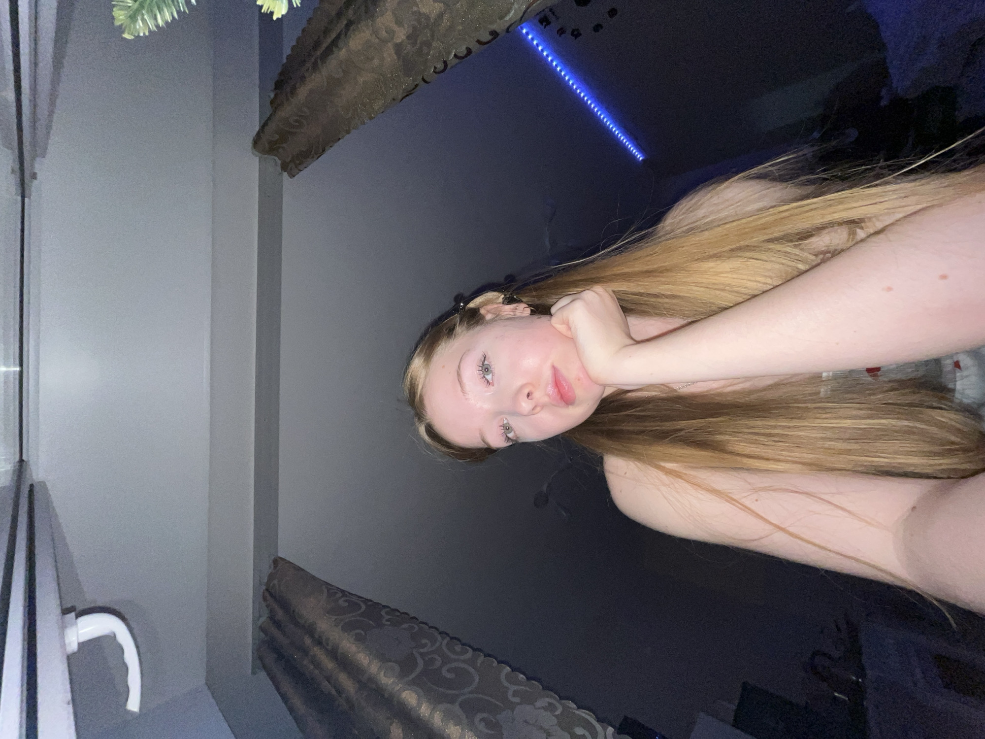 A fair-skinned person with long, straight blonde hair is reclining, head resting on their right hand, in what appears to be an indoor setting. The background is largely dark, with a faint linear blue light source visible in the upper part of the frame. A curtain hangs in the upper-left corner of the image, and a window with a handle is visible on the left side. There is insufficient information to establish a location in Chaikovsky, Russia. The person's expression is neutral, and their gaze is directed towards the camera.