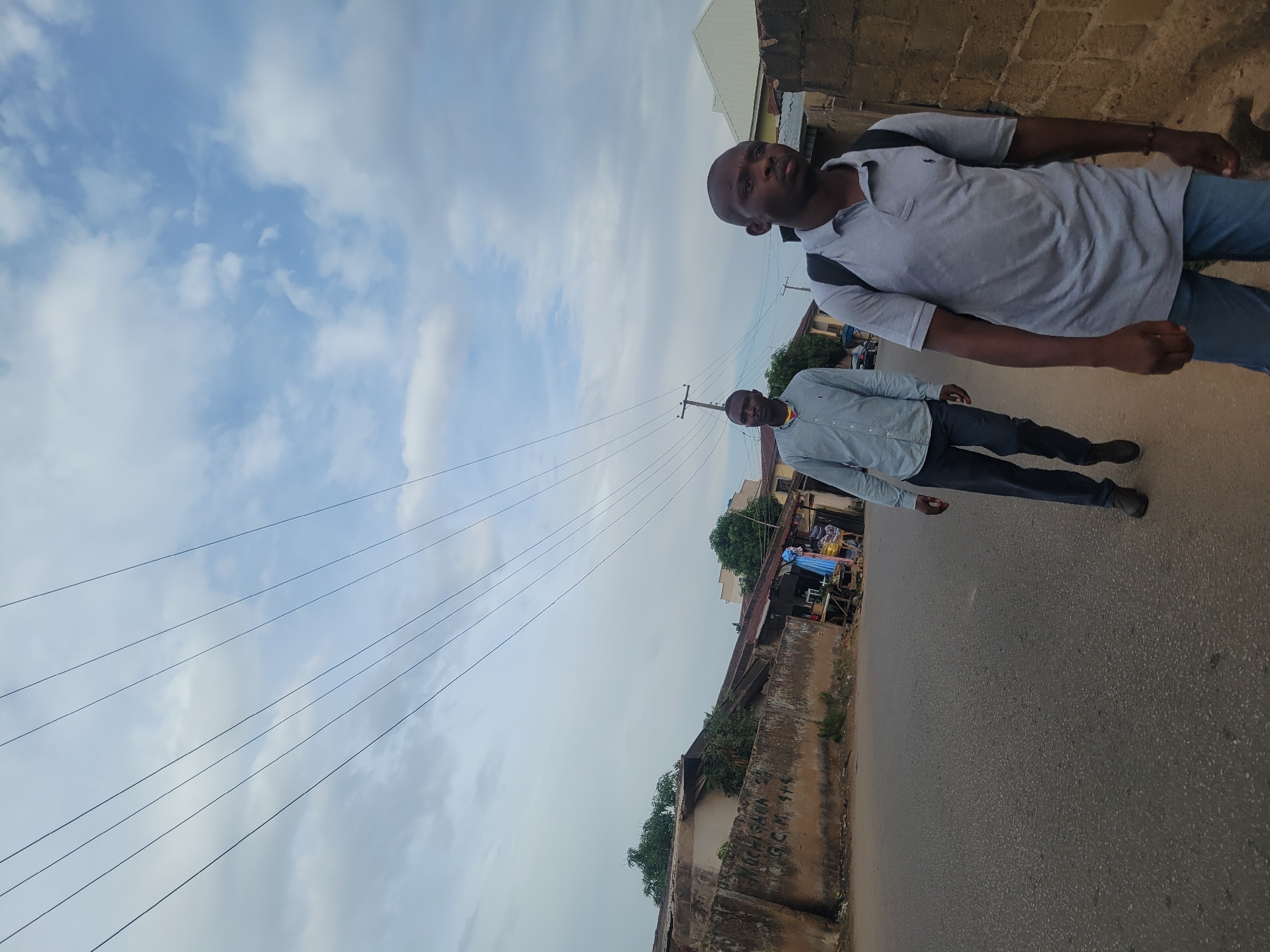 The image features two men standing on a paved street in an urban or semi-urban environment in Jos, Nigeria, under an overcast sky during daytime. The perspective is tilted, making the street and subjects appear at an angle.
The man in the foreground, positioned on the right, wears a light grey short-sleeved polo shirt, dark blue jeans, and a black backpack. He has short hair and is looking directly towards the camera. Further down the street, slightly behind him and more towards the center of the frame, stands another man. He is dressed in a light-colored, possibly checkered, long-sleeved button-down shirt and dark trousers, also looking at the camera.
Numerous dark utility lines stretch across the sky, originating from poles partially visible along the street. Buildings, some with corrugated metal roofs, line both sides of the road, interspersed with green foliage. A weathered concrete wall in the lower left foreground exhibits some faded, indistinct writing, which appears to include numbers like "999" or similar characters. The sky is predominantly cloudy with hints of blue showing through. The men appear to be posing for a photograph.