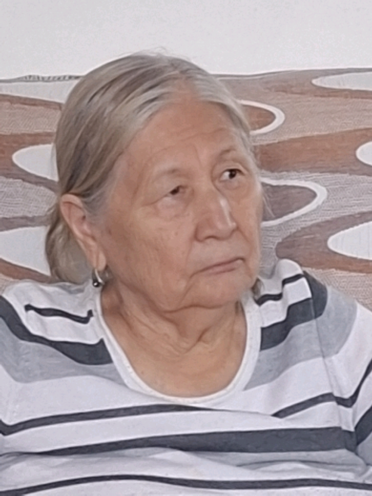 An elderly woman with grey hair pulled back is seated, facing right, wearing a striped shirt. She has a hoop earring in her left ear. The background shows a patterned surface, possibly a sofa or wall. The image is a close-up shot focused on the woman's face and upper body. There is no discernible location within the city of Almaty, Kazakhstan.