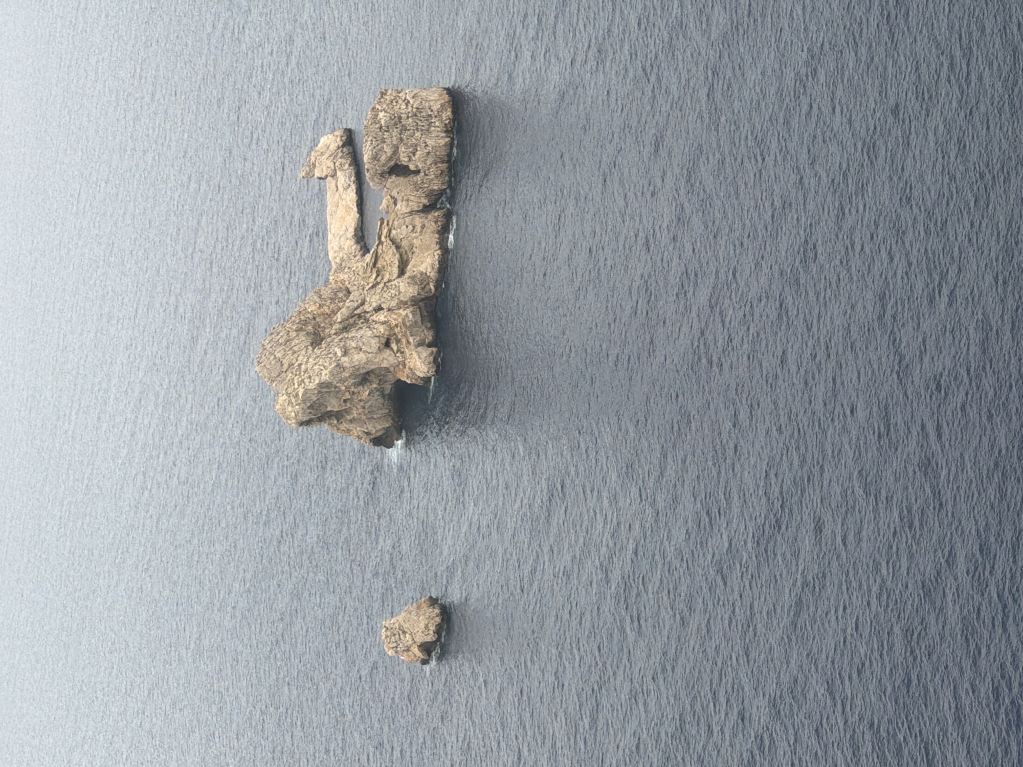 The image displays an aerial view of a rugged rock formation emerging from a dark, choppy body of water. The rock is predominantly tan and brown, with visible textures of erosion and uneven surfaces. It appears to be a collection of islands or a large island broken into sections, with some smaller rocks in the foreground. The water is a deep, muted blue-gray and shows ripples and small waves, suggesting a moderate level of movement. The sky above is overcast and a pale gray, contributing to the muted color palette of the scene. There are no people or man-made structures visible in the image.
The rock formation is located off the coast of Sant Antoni de Portmany, Spain, specifically near the Cap Blanc lighthouse.