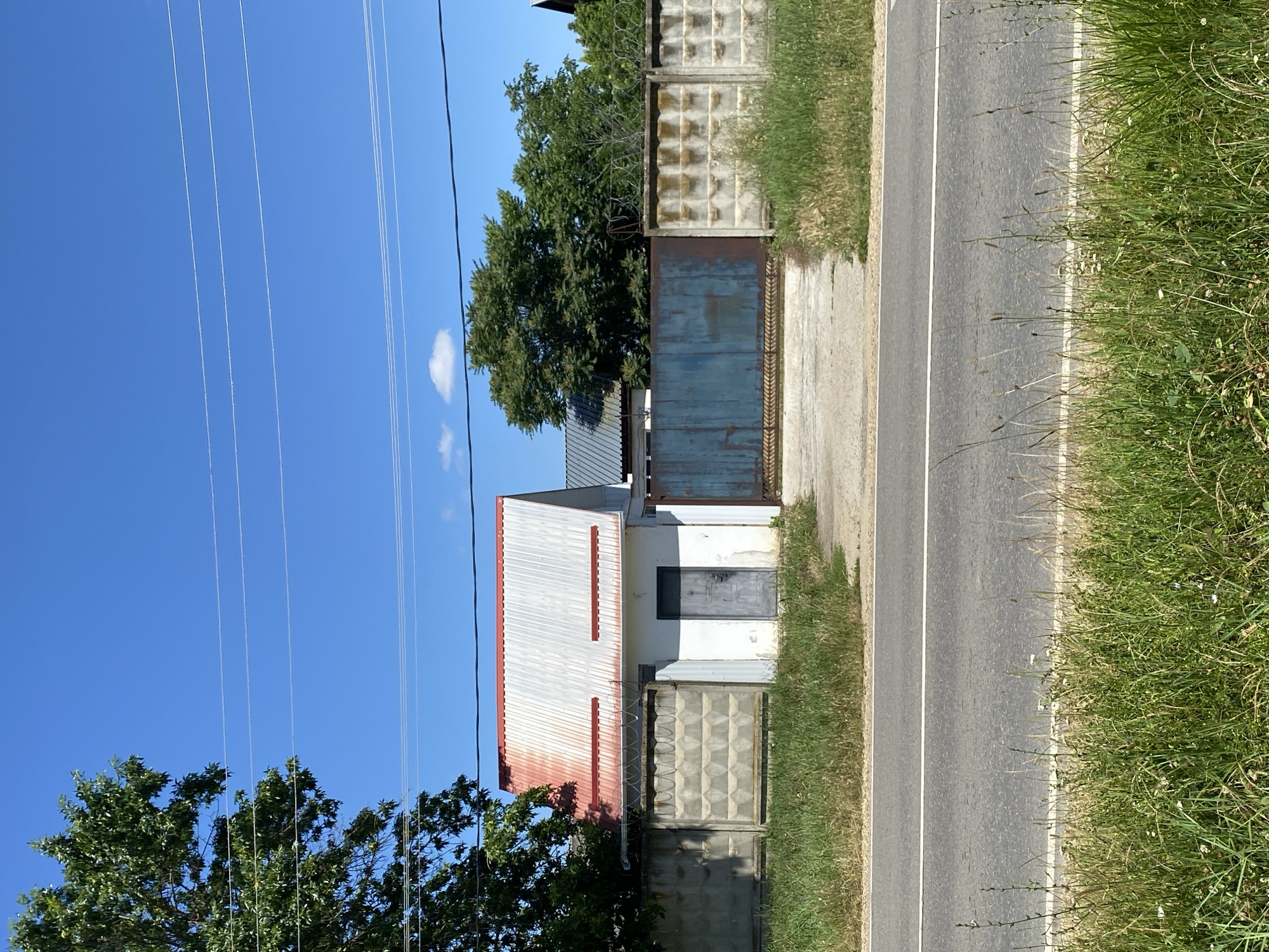 The image captures a roadside scene in an outdoor environment, likely on a sunny day with a clear blue sky. A grey asphalt road with a white dividing line runs horizontally across the lower portion of the frame. On the opposite side of the road, a patch of green grass and some wild vegetation borders a concrete fence. The fence is constructed from pre-cast concrete panels with a repeating geometric pattern.
Behind the fence, a small white building with a corrugated metal roof, partially painted red, is visible. The building has a single grey door. To the right of this building, a large, weathered, rusty blue metal gate stands, also partially obscured by the concrete fence. Further behind the gate and fence, lush green trees are prominent. Overhead, several power lines stretch across the sky. The overall impression is of a rural or semi-urban setting, possibly the outskirts of a city like Zapadny, Russia, given the architectural style and vegetation. No people are present in the image.