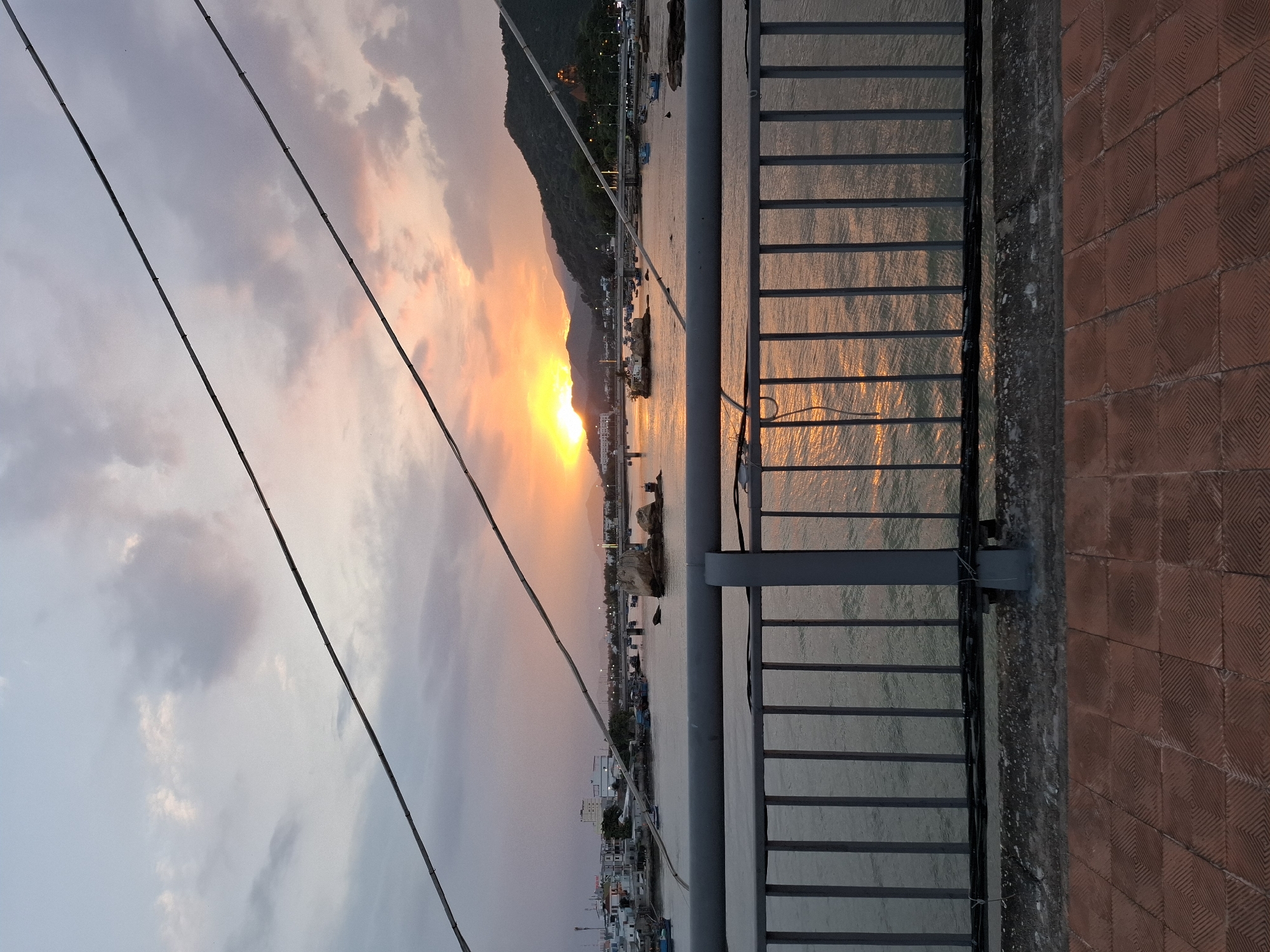 The image shows a sunset over a river in Nha Trang Ward, Vietnam. The sun is low on the horizon, casting a warm glow on the sky and water. Several boats are anchored in the river, and a few buildings are visible on the shore. The foreground of the image features a bridge railing and a tiled walkway. The scene appears peaceful and serene.