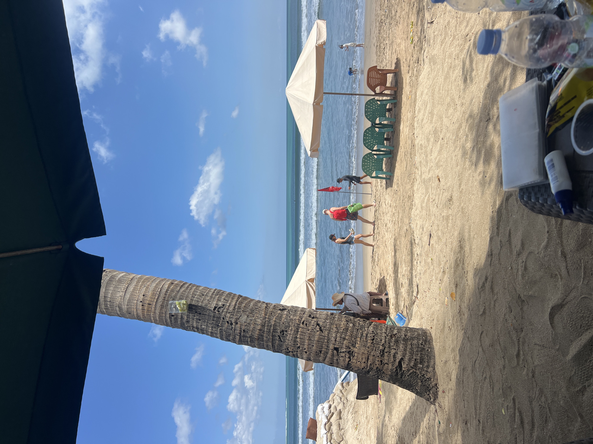 In Kuta, Indonesia, a sandy beach is depicted under a bright blue sky with scattered white clouds. A large palm tree trunk stands in the foreground, angled towards the left, with a clear plastic container holding green items attached to it. The middle ground features the ocean with moderate waves, and several people are visible. Two individuals are playing what appears to be a game on the sand, one holding a red flag. Further back, near a light-colored beach umbrella, several green plastic chairs are arranged, with one brown plastic chair to the right. Other beachgoers are scattered along the shoreline, with one person wading in the water and another near the umbrella. Sandbags are piled up on the left side of the frame, with a black chair resting on them. In the foreground, a dark woven surface holds two plastic water bottles, a white spray bottle, a clear plastic cup, and a clear plastic pouch containing white papers.