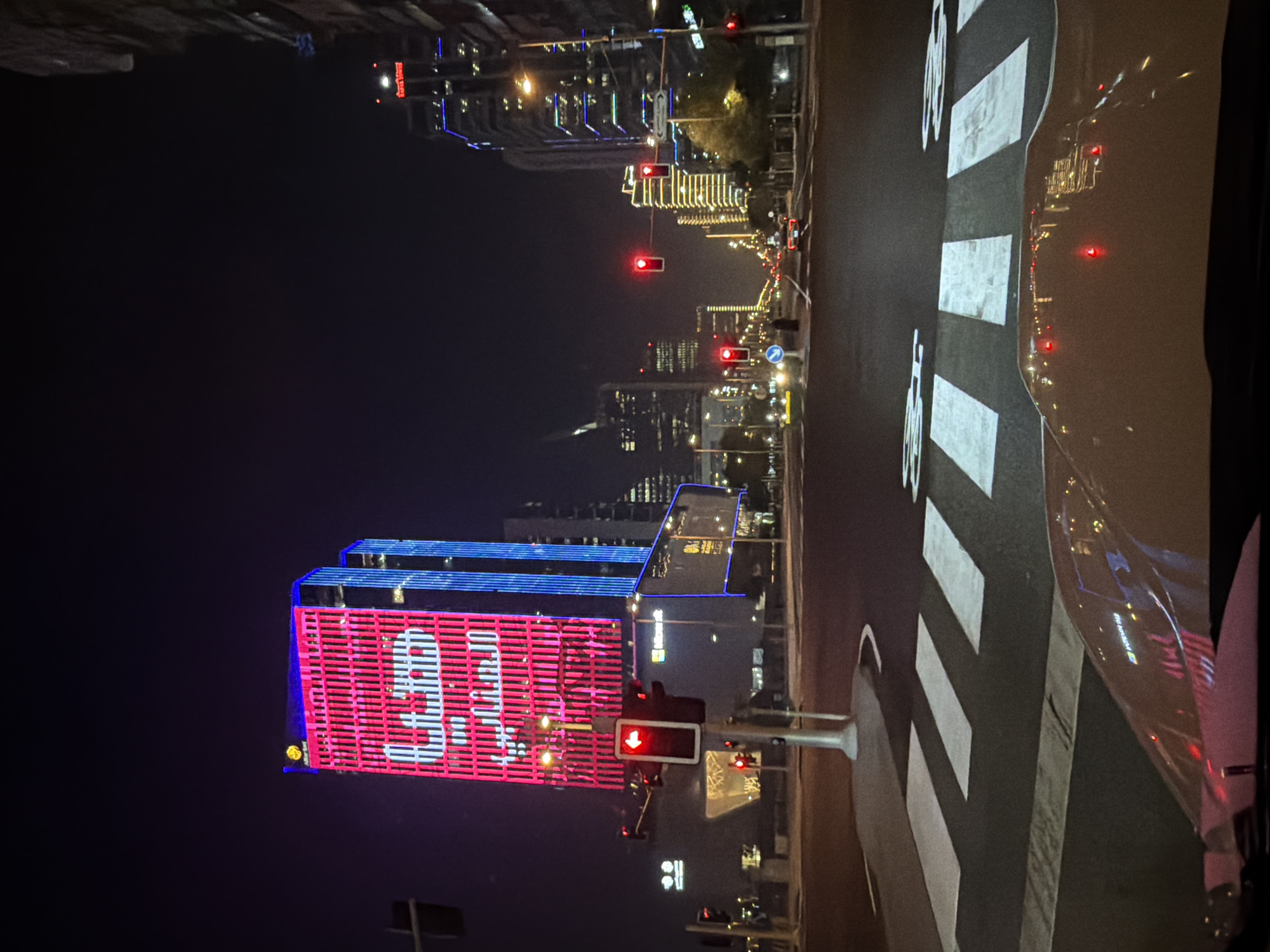 The image depicts a nighttime urban scene. A pedestrian crossing with white stripes and bicycle symbols is visible in the foreground, illuminated by streetlights and reflections from nearby buildings. To the left, a building is brightly lit with neon signs. The sign displays large white numbers, possibly indicating a time or a promotional message, set against a pink and blue striped background. Further down the street, other buildings are illuminated with various colored lights, creating a vibrant city skyline. Traffic lights are visible, with at least one showing a red light, indicating a stop. The scene suggests a busy city environment at night, with activity on the street and in the surrounding buildings. The overall impression is one of a modern, well-lit urban landscape. There is no clear indication of people, weather, or specific location cues beyond the urban setting.