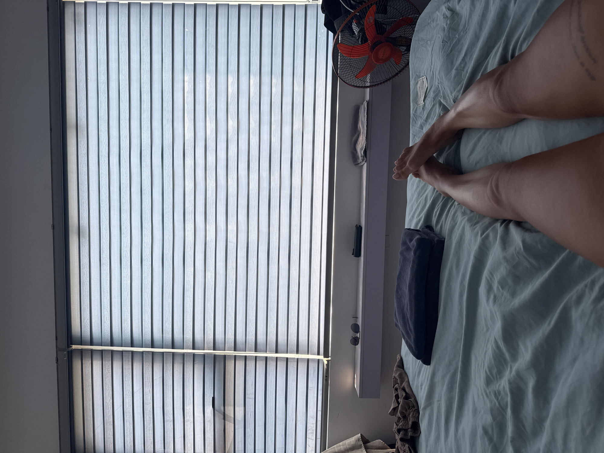 A person lies on a bed covered with a light-blue sheet. The person's legs, from the thighs down, are visible; the feet are crossed at the ankles. A fan is visible near the head of the bed. Beside the bed is a narrow shelf with various items, including a pair of sunglasses. The window has closed vertical blinds. It is hard to say for certain whether the picture was taken in the city of Đà Nẵng, Vietnam.