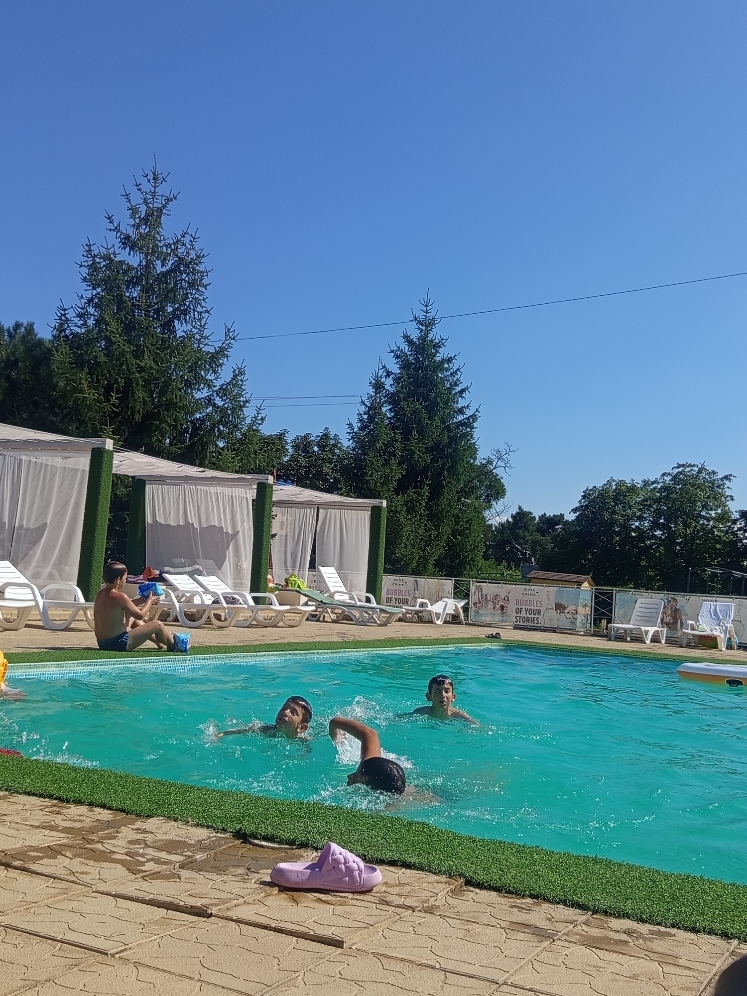 The image depicts a sunny day at an outdoor swimming pool area, likely located in or near Molovata Nouă, Moldova. In the foreground, the turquoise water of the pool is visible with several young males swimming. One person, appearing to be a boy, is in the center of the pool, looking towards the camera and swimming with his arms outstretched. Another person, also appearing to be a boy, is swimming further back in the pool, with only his head and shoulders visible above the water. To the left of the pool, a young male is sitting on the edge, dressed in swim trunks and holding a blue object, possibly a water gun. He is seated on artificial turf that surrounds the pool. In the background, several white lounge chairs and cabanas with white drapes are lined up on a paved area. Trees and dense foliage surround the pool area. A banner with text and images is visible in the background behind some of the lounge chairs. The sky is clear and blue. A single pink flip-flop lies on the paved ground to the right of the pool.