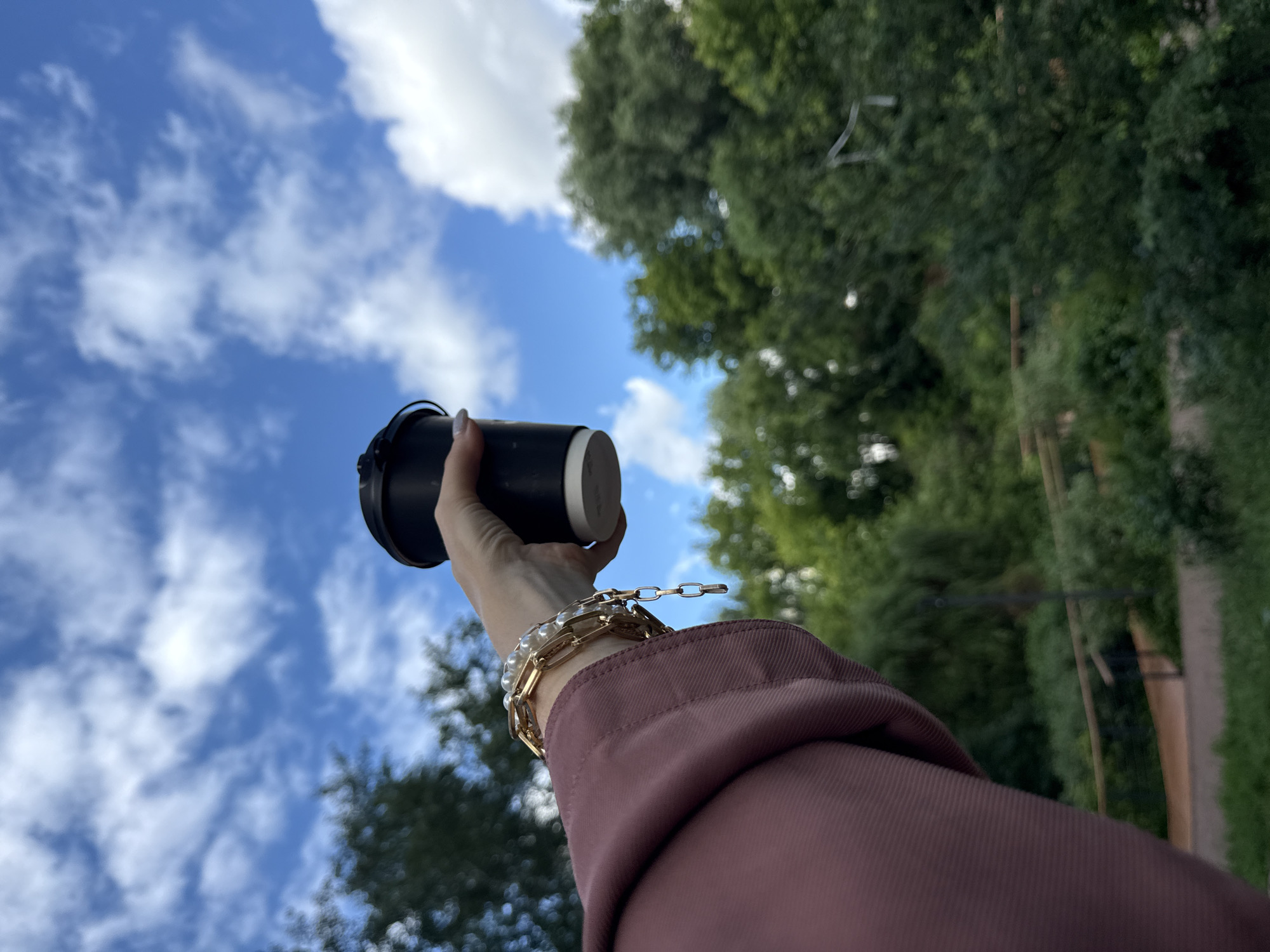 A hand, adorned with pearl and gold bracelets, holds a black travel mug against a backdrop of blue sky with scattered white clouds and lush green trees. The shot is taken from a low angle, emphasizing the sky and the hand extending upwards. The person is wearing a muted rose-colored jacket. The lighting suggests it is daytime, likely sunny given the bright sky. The background trees are dense and full, indicating a warm season. While the exact location is not explicitly visible, the presence of trees and a clear sky are common in outdoor settings. There is no visible text in the image.