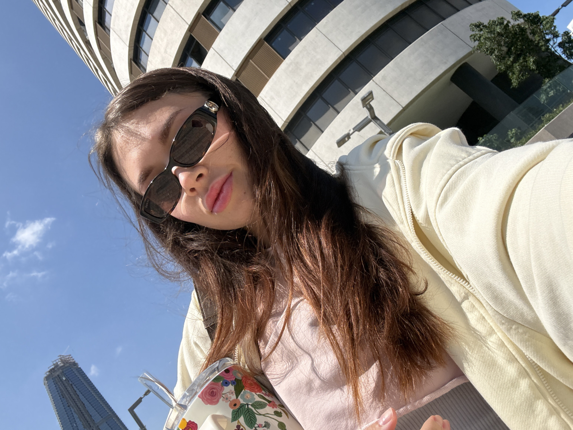 A woman with brown hair and sunglasses is taking a selfie from a low angle. She is wearing a light-colored jacket and a pink top. She is holding a cup decorated with floral patterns. In the background, there is a curved modern building with horizontal windows, and in the distance, there is a skyscraper under construction. The clear blue sky indicates a sunny day. Based on the architecture and the presence of the modern skyscraper, the location is likely in Tel Aviv, Israel.