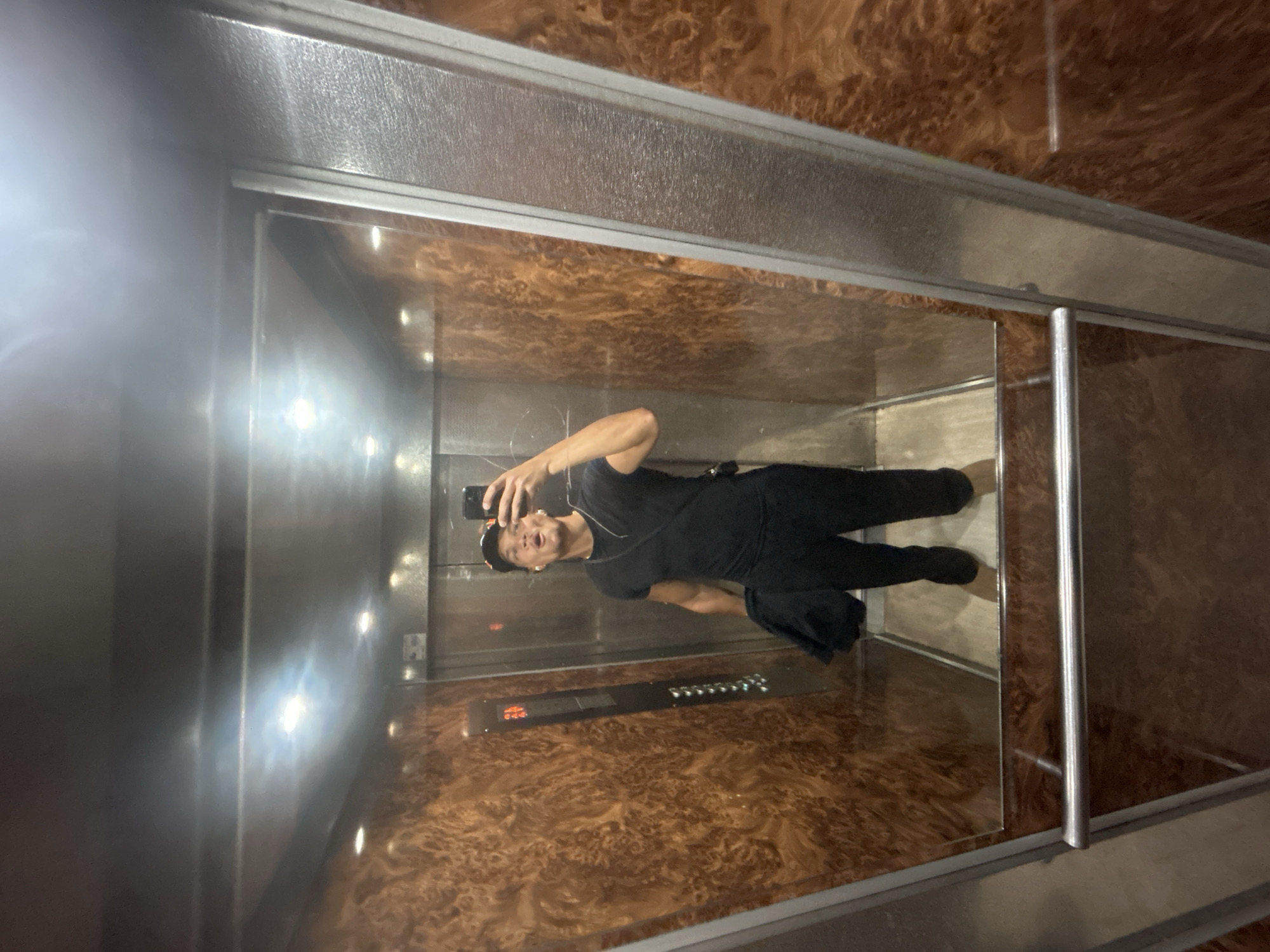 The image captures a selfie taken by a young man inside an elevator. The man is reflected in a highly polished, mirror-like surface, likely the elevator door or a mirrored wall. He is dressed casually in a black t-shirt, black trousers, and a black baseball cap worn backward. With his right hand, he holds a smartphone pointed towards the reflection, his mouth slightly open in an expression for the photograph. He also wears a thin necklace and an earring.
The elevator interior showcases a mix of materials. The wall to the viewer's left (which appears as the right wall in the man's reflection) is made of brushed stainless steel. The opposite wall, where the selfie is taken, is a reflective, mirrored surface. On this mirrored surface, an elevator control panel is visible, featuring multiple buttons and a digital display. The display clearly shows the number "3," indicating the current floor. A silver handrail is also present along this wall.
The back wall and ceiling of the elevator are covered in a distinctive, dark brown, burled wood-like paneling, providing a warm contrast to the metallic elements. Artificial lighting, likely from embedded fixtures, illuminates the space and creates bright reflections on the shiny surfaces. The overall setting is a modern and clean elevator, consistent with a building in Noisy-le-Grand, France.
