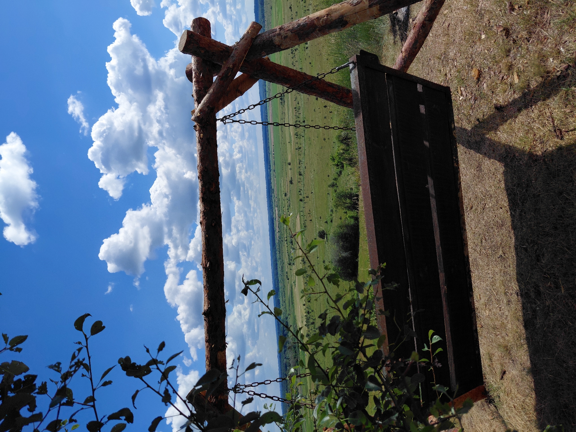 This is a photograph of a wooden swing set situated on a grassy hill overlooking a rural landscape under a partly cloudy sky. The swing itself is made of dark wood and hangs from thick chains attached to a rough-hewn wooden frame. In the foreground, the edge of the swing and some branches with green leaves are visible, partially obscuring the view. The background features a vast expanse of green fields dotted with trees and bushes, stretching towards a distant horizon. The sky above is a vibrant blue, punctuated by fluffy white cumulus clouds. The lighting suggests it is daytime, possibly late morning or early afternoon, with strong sunlight casting shadows. The location is identified as Ustyvytsia, Ukraine. There are no people visible in the image, and no actions or interactions are depicted.