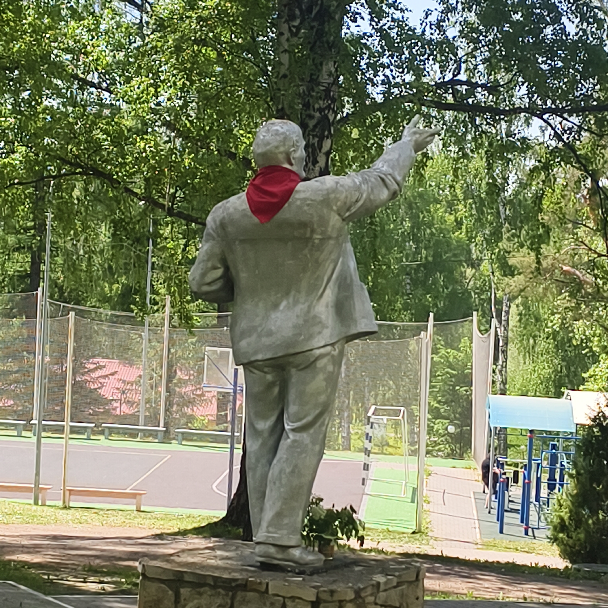 The image depicts a statue of a man with a red scarf around his neck, viewed from the back. The statue is set on a stone base in a park. A birch tree stands behind the statue, partially obscuring it. In the background, there is a sports court with benches and netting, a playground structure, and lush greenery. The location is Варзи-Ятчи, Russia.