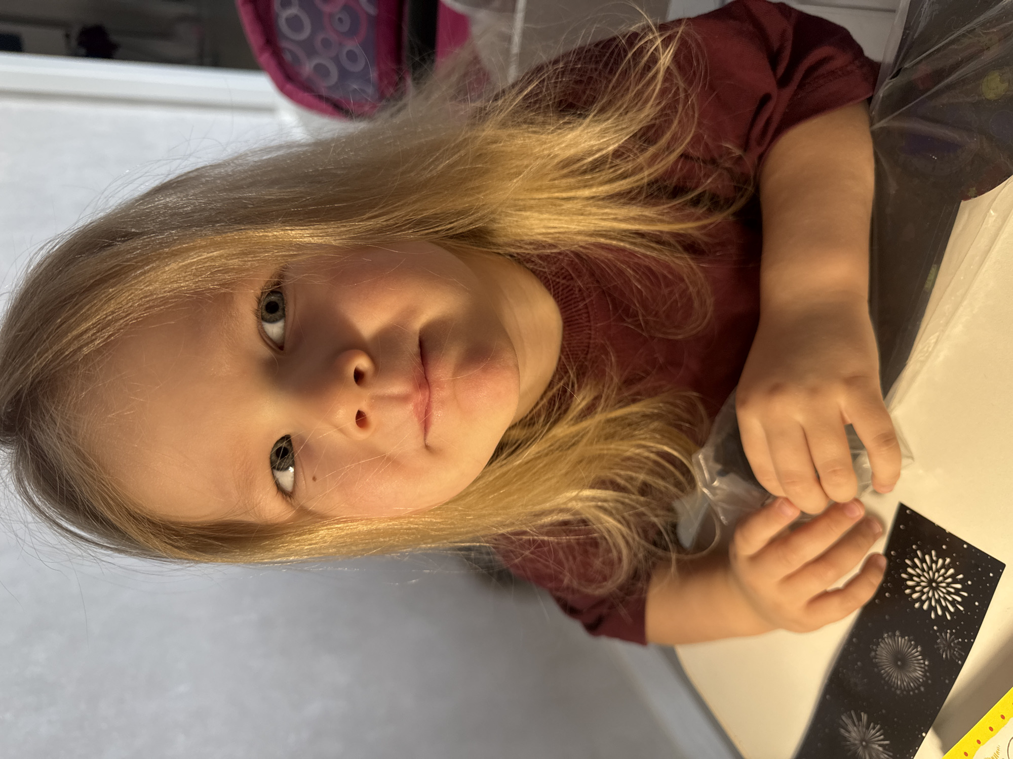 A young, fair-skinned child with long, light blonde hair is indoors, wearing a burgundy t-shirt, and sitting at a white table. The child is looking directly at the camera. The child's right hand is holding a clear plastic bag. On the table to the child's right, there's a black card with white firework-like designs, and a portion of a yellow object. The location is Gornyi (Dvurechenskoe S/P), Russia.
