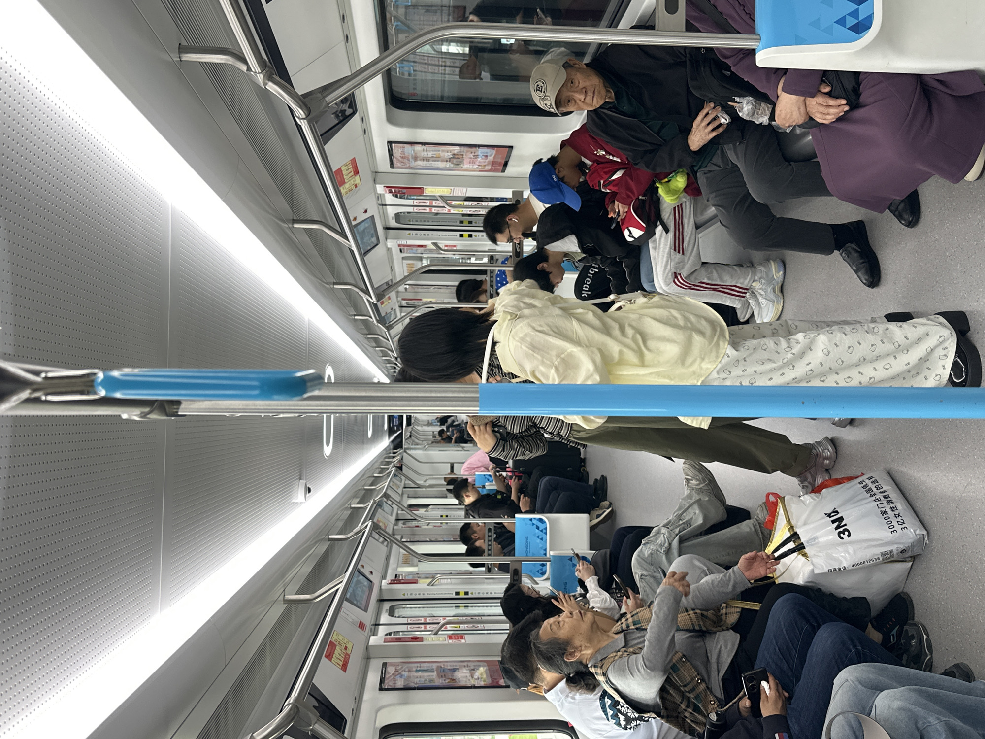 The image captures the interior of a subway train car in Jiangjin, China. The carriage is filled with passengers, many of whom are seated. An elderly man wearing a white baseball cap is visible towards the top right. Some passengers are using their cell phones. A large white shopping bag with Chinese text printed on it rests on the floor. The train's interior features handrails and advertisements on the walls. The lighting is bright.