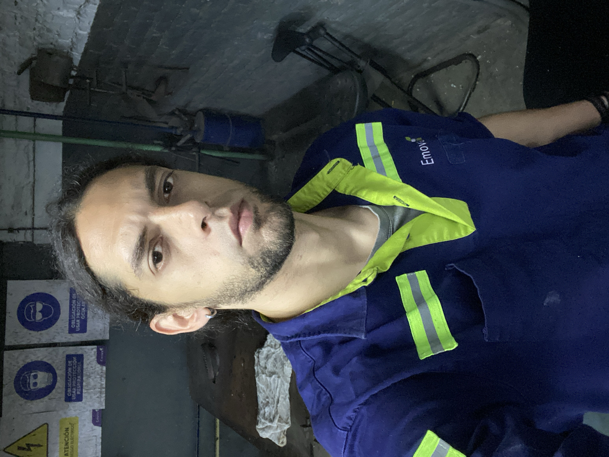 A selfie shows a man with long dark hair and a short beard. He wears a dark blue work shirt with neon green trim and the logo "Emove" is visible on the upper right chest. He has an earring in his left ear. Behind him is a wall with safety signs written in Spanish and a high-voltage electrical hazard sign. Other details include some exposed pipes and a tool bench in the background. Given the presence of Spanish signage and Emove branding, it is possible this scene is located in Buenos Aires, Argentina.
