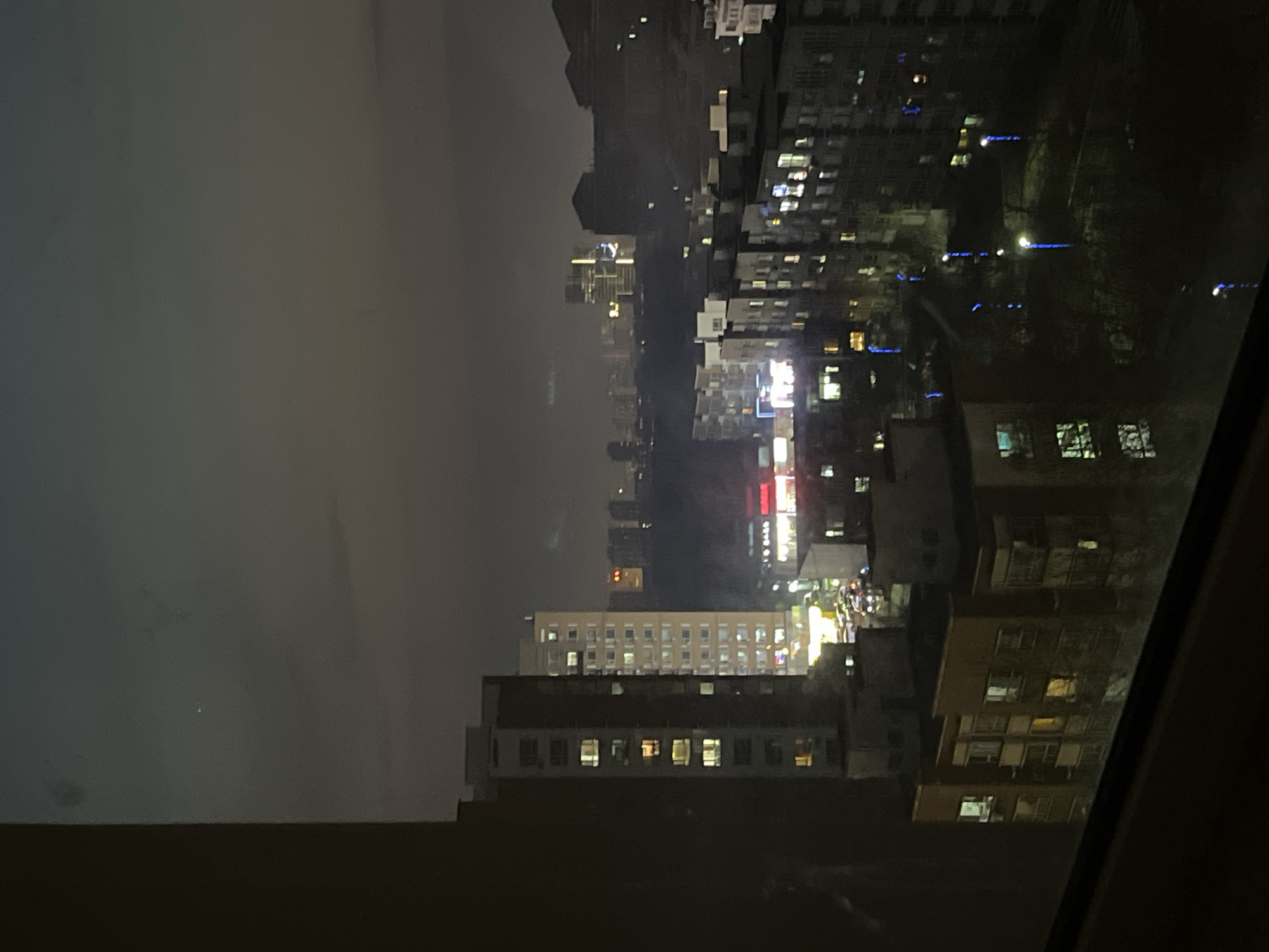 The image shows a night view of Changchun City, China, from an elevated position, possibly a high-rise building. The scene is dimly lit with city lights illuminating various buildings. Skyscrapers and apartment buildings are visible in the distance, shrouded in a haze or fog. Some buildings have brightly lit windows, while others are dark. There is some reflection on the window or surface where the picture has been taken. The overall environment is urban, with the typical characteristics of a nighttime cityscape.