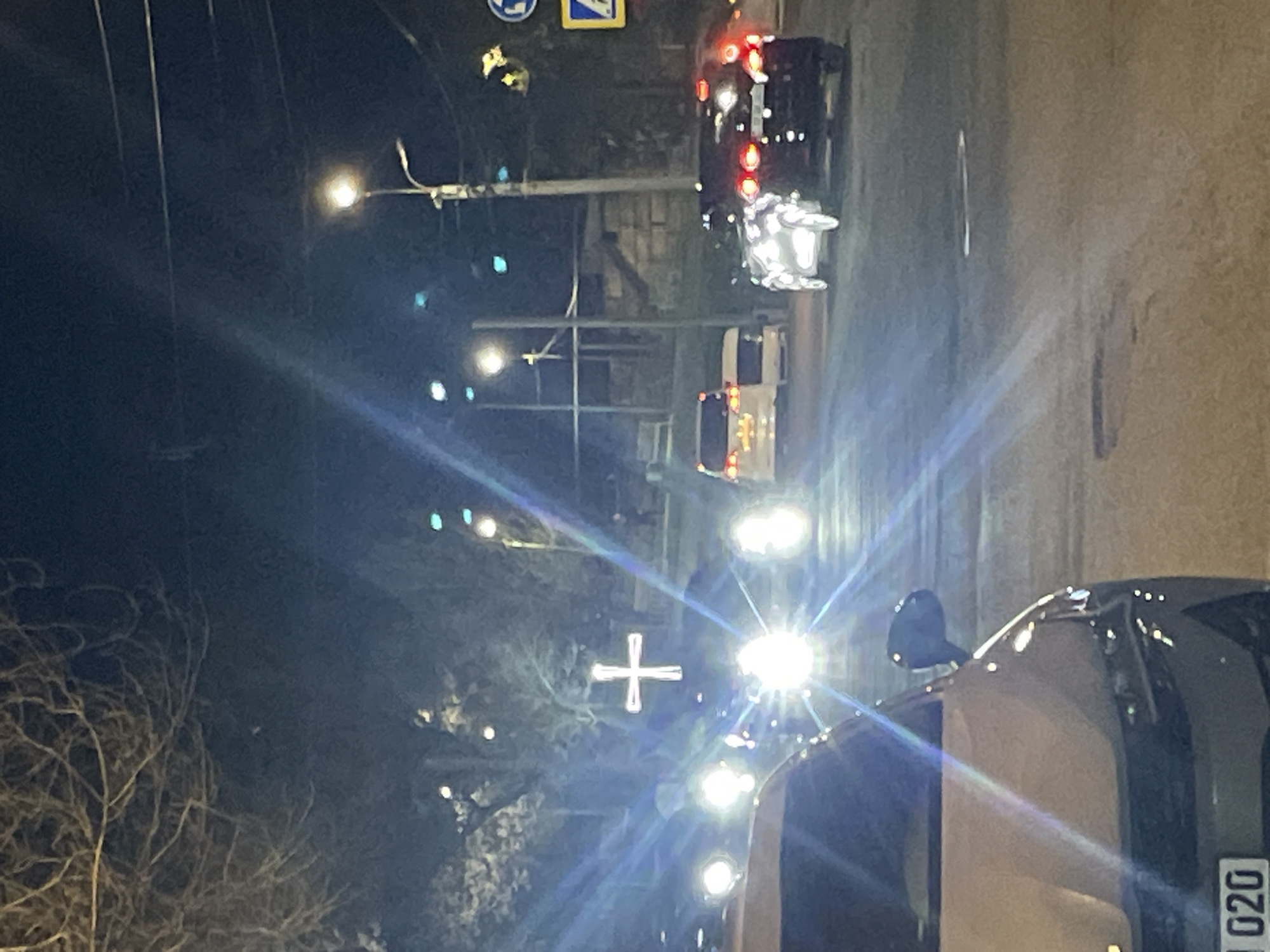 The image depicts a nighttime street scene in Yerevan, Armenia. Several cars with illuminated headlights and taillights are visible on the road. Streetlights are present, casting light on the surroundings. In the background, a Christian cross is illuminated. Branches from a tree can be seen on the left side of the frame. A road sign is visible on the upper left side of the image. A car is parked or driving in the foreground with the license plate number ending in "020". The scene is lit by artificial lights and vehicle headlights.