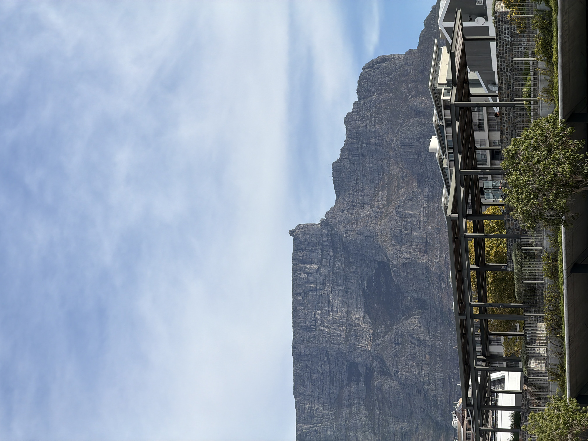 The image shows Table Mountain in Cape Town, South Africa. The mountain is seen on the right side of the frame, adjacent to buildings with a metal structure. The sky is visible on the left side, with white clouds against a light blue background.