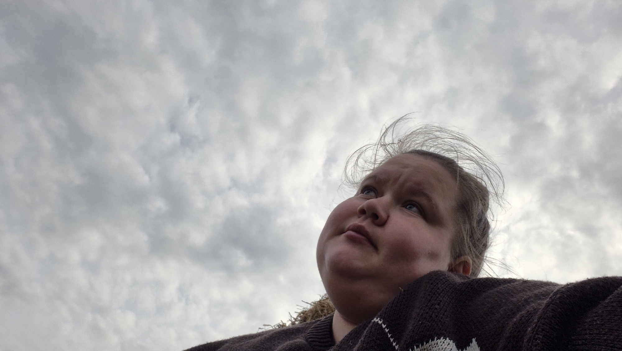 A low-angle outdoor shot captures a person, likely a woman, from the chest up, positioned in the lower right portion of the frame, looking upwards and to the left. The dominant feature of the image is the vast, cloudy sky, which fills approximately three-quarters of the frame. The clouds are uniformly grey and white, indicating an overcast day.
The person has light-colored hair, some strands of which appear windswept around their head. Their face is seen in profile, with their blue eyes directed towards the upper left. Their expression appears pensive or observant as they gaze at the sky. They are wearing a dark brown, possibly knitted, garment that covers their shoulders and part of their arm, with a white, geometric pattern visible on the fabric in the lower right corner. The angle emphasizes the underside of their chin and jawline. No specific location cues are visible within the image to identify Дойная, Russia, beyond the general outdoor setting with an expansive sky. There is no visible text in the image.