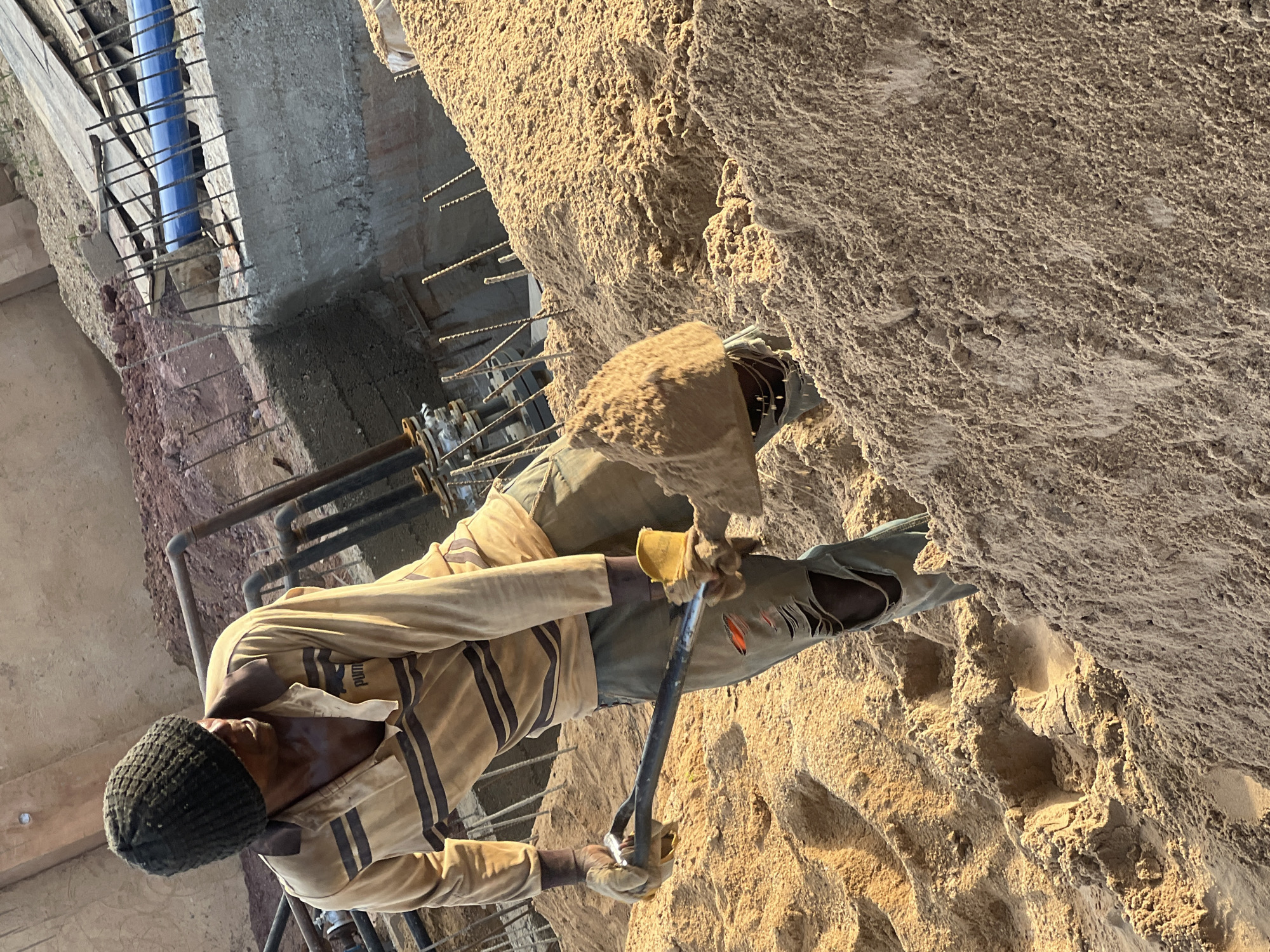 An outdoor, eye-level shot captures a man engaged in manual labor at what appears to be a construction or excavation site in Jos, Nigeria. The man, seen from the waist up and turned slightly to his right, is wearing a striped beige and dark-brown long-sleeved shirt, dark pants, a dark knit beanie, and light-colored work gloves. Faint text reading "PUMA" is visible on his shirt. He is holding a heavy metal tool, possibly a pickaxe or pry bar, with both gloved hands, positioned near a textured, sand-colored surface that looks like natural rock formation or compacted soil.
The environment is characterized by this rough, sandy-textured material, which forms a large wall-like structure to the man's right and extends across the ground. In the background, exposed concrete structures with protruding rebar (metal reinforcement rods) are visible, along with sections of metal piping and a blue pipe, indicating ongoing construction or earthwork. The scene is brightly lit by natural sunlight, suggesting daytime hours. The man appears focused on his task, working with the heavy tool in the dusty, dry surroundings.